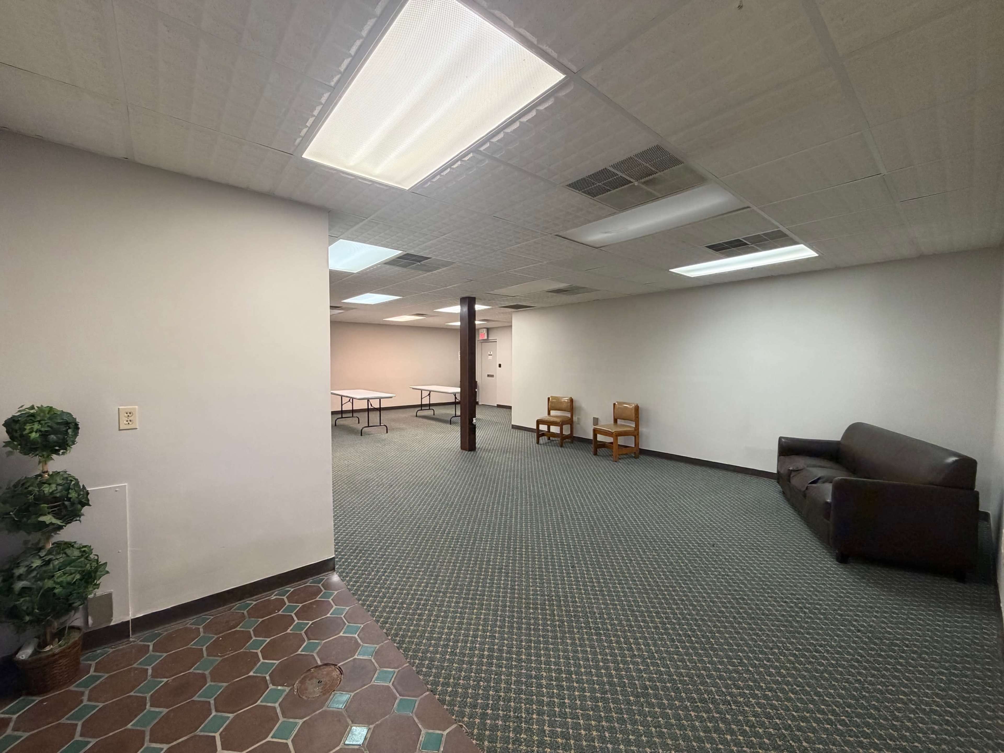 The image shows an empty room with a carpeted floor, two wooden chairs, a couch, and a few tables set up in the back.