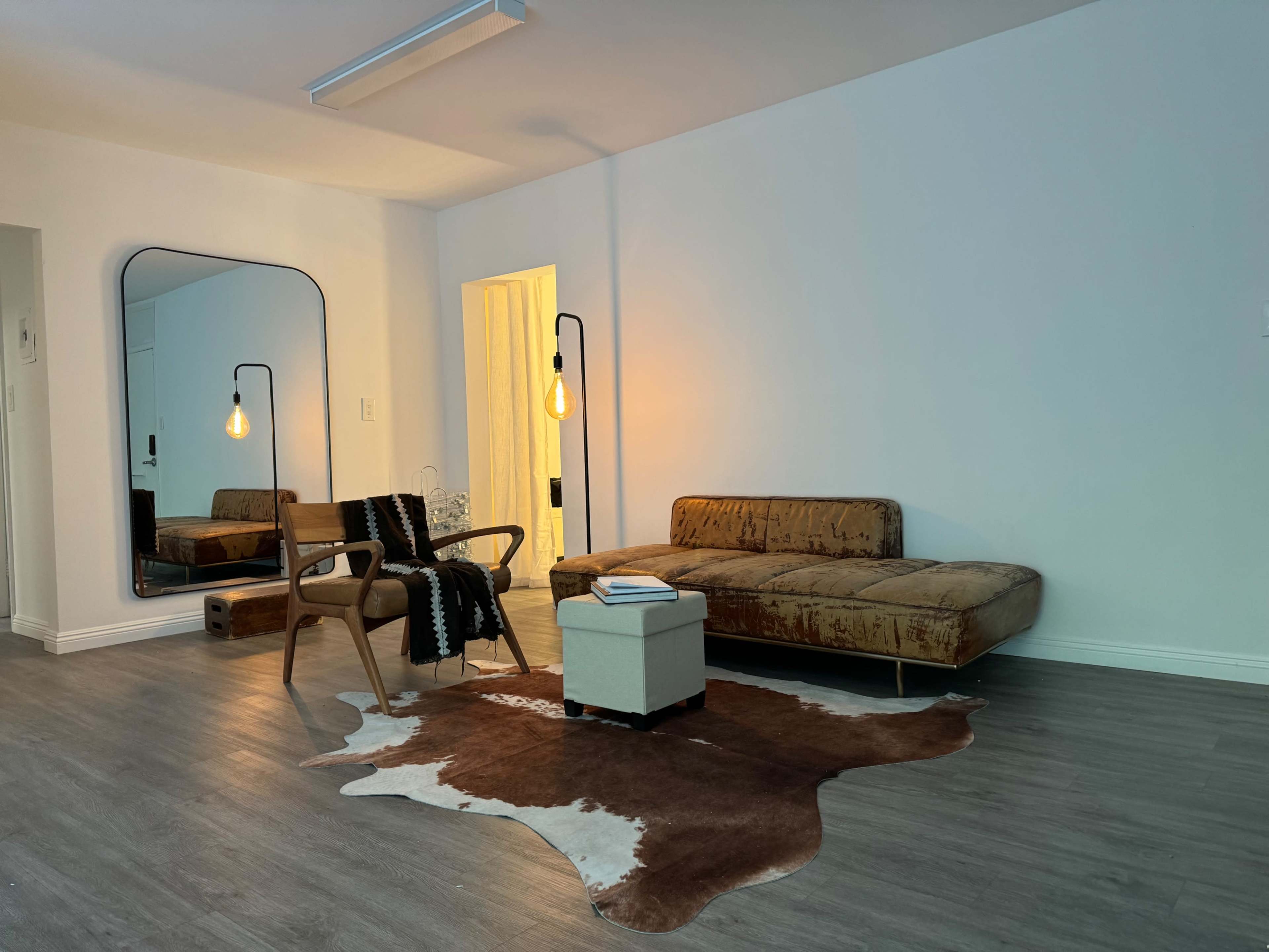 A modern living room features a brown sofa, a wooden chair, a light fixture, a cowhide rug, and a small table.