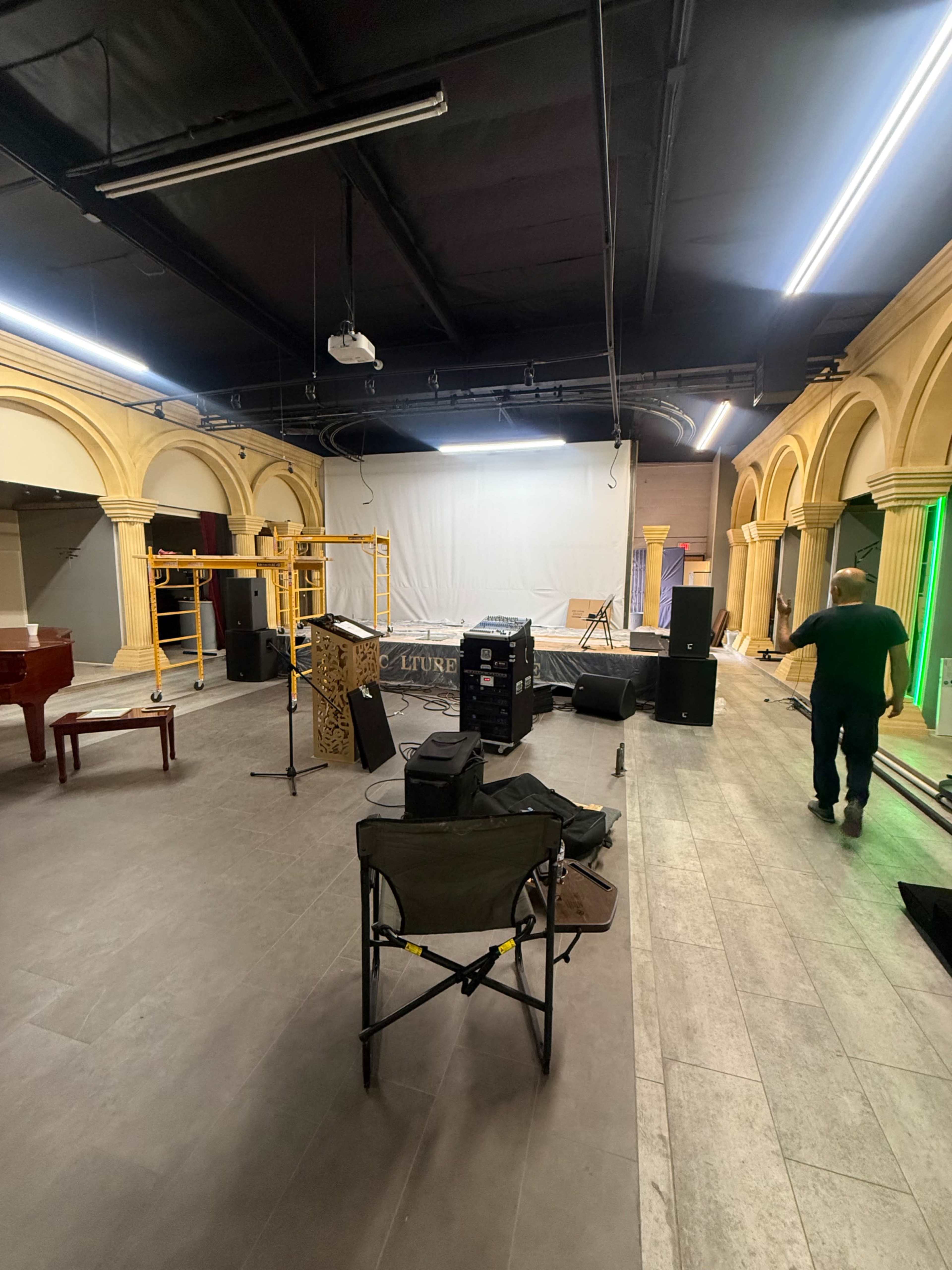 The image shows an empty performance space with a stage in the background, various audio equipment, and a man walking towards the right.