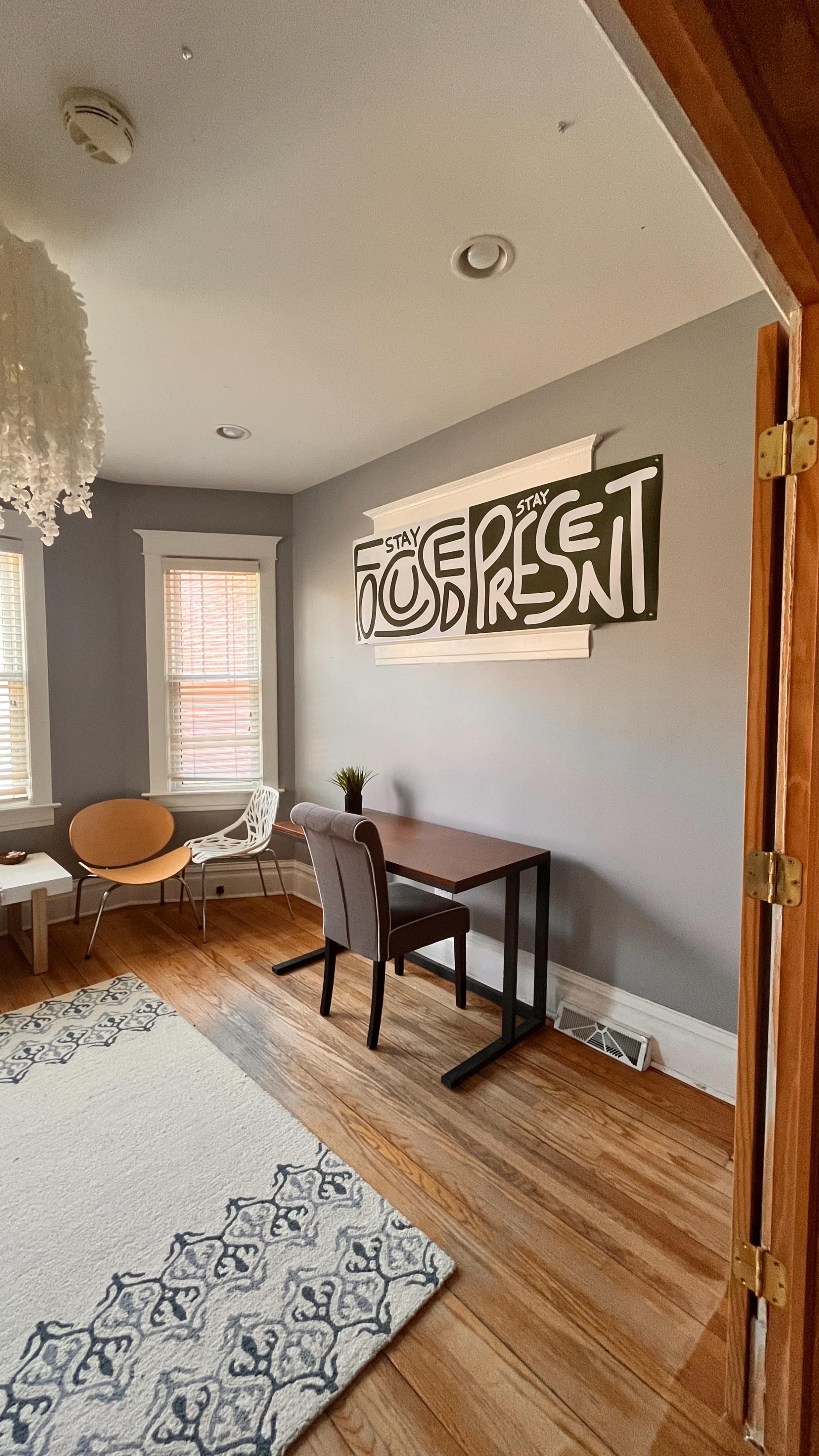 The image shows a cozy interior room with hardwood flooring, featuring a table and a chair, along with a decorative wall that has the phrase "STAY FOCUSED STAY PRESENT" displayed prominently.