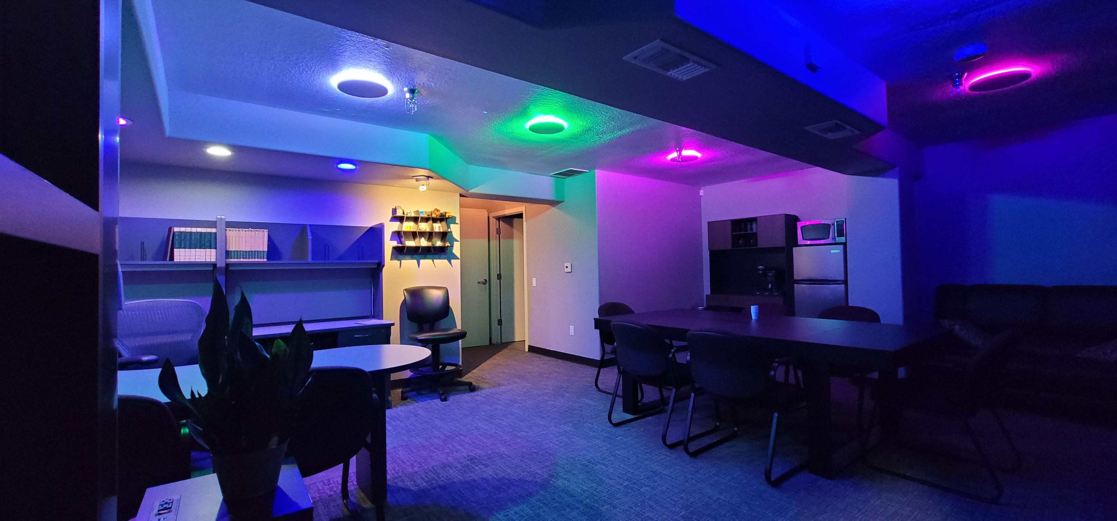 The image shows a modern, dimly lit room with colorful LED lights, a table surrounded by chairs, and a kitchenette in the background.