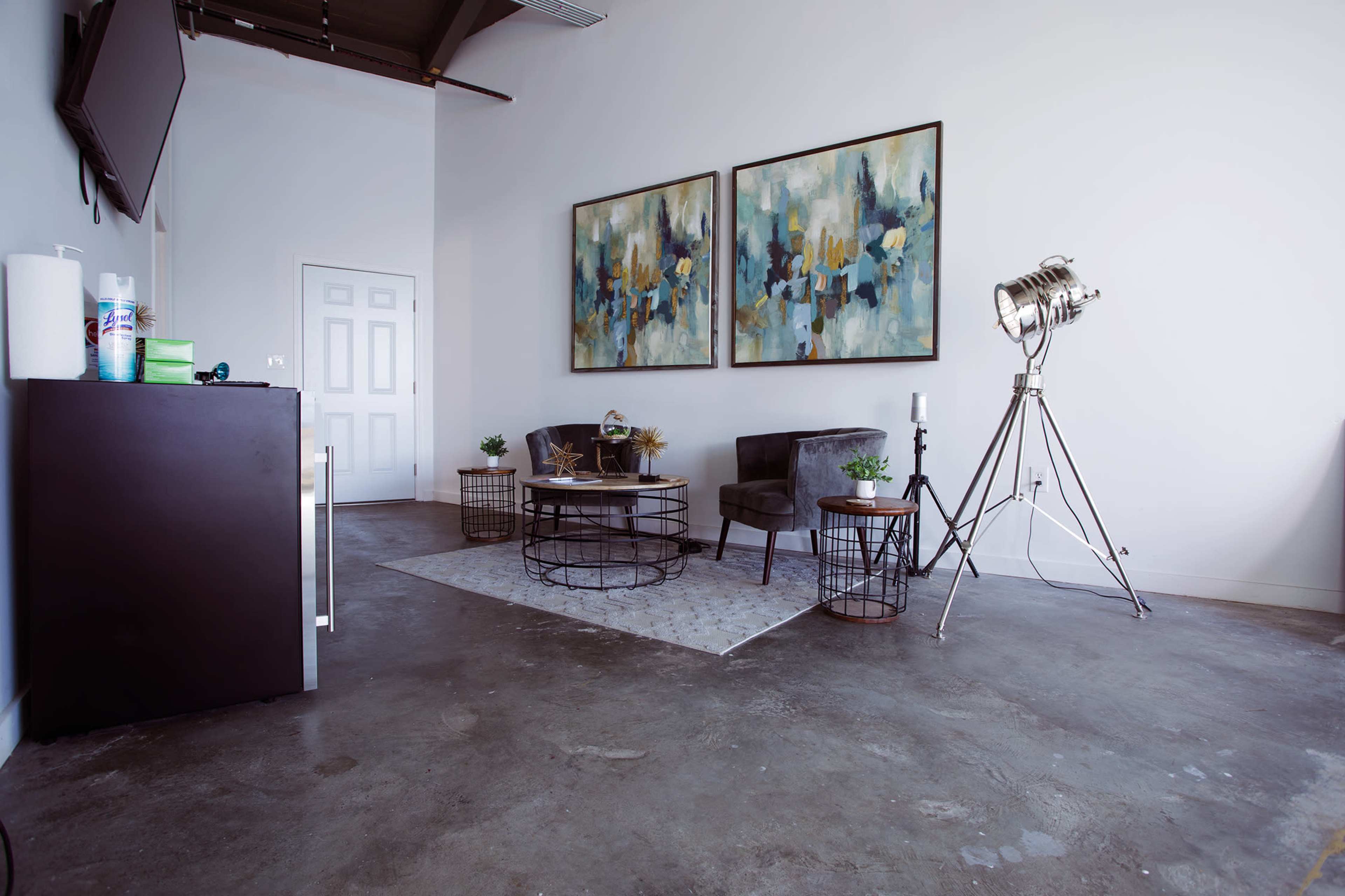 The image depicts a modern interior space featuring two upholstered chairs, a coffee table, two wire side tables, and abstract artwork on the walls, with a spotlight lamp and a large door in the background.