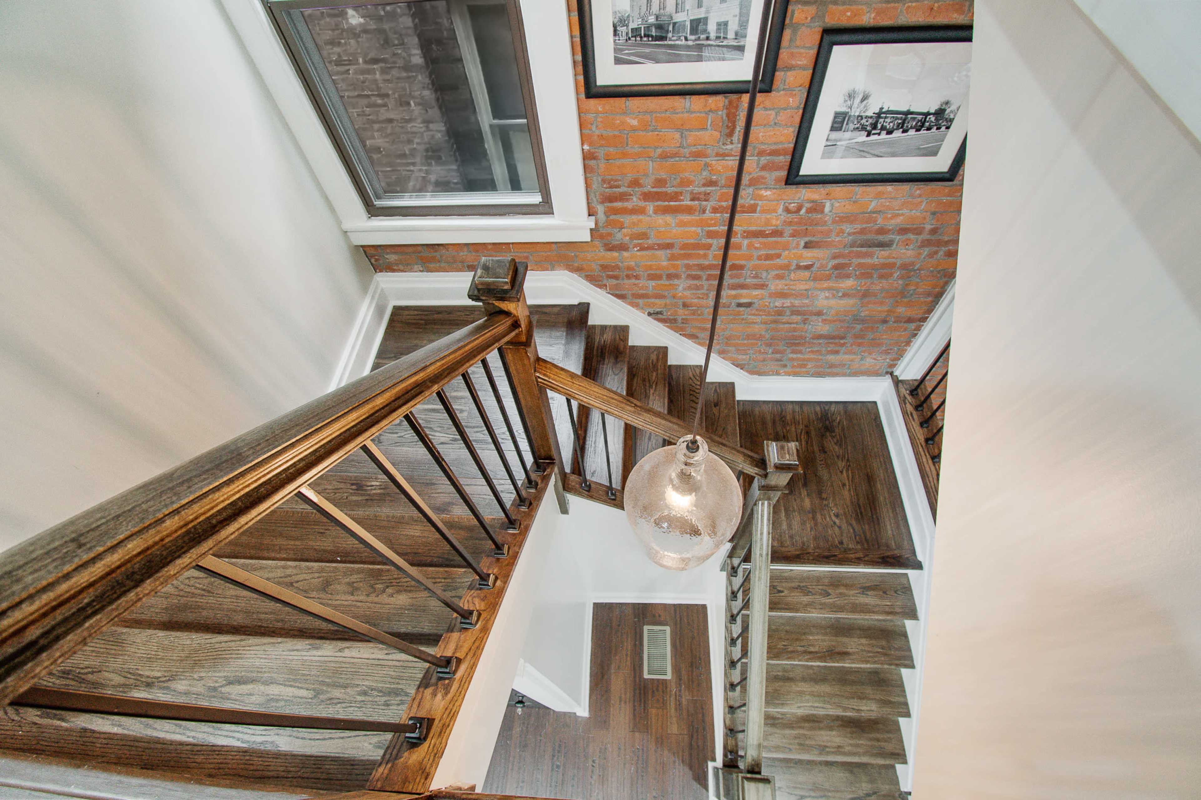 A wooden staircase with a metal railing leads downwards, accompanied by framed photographs on the wall and a hanging light fixture above.