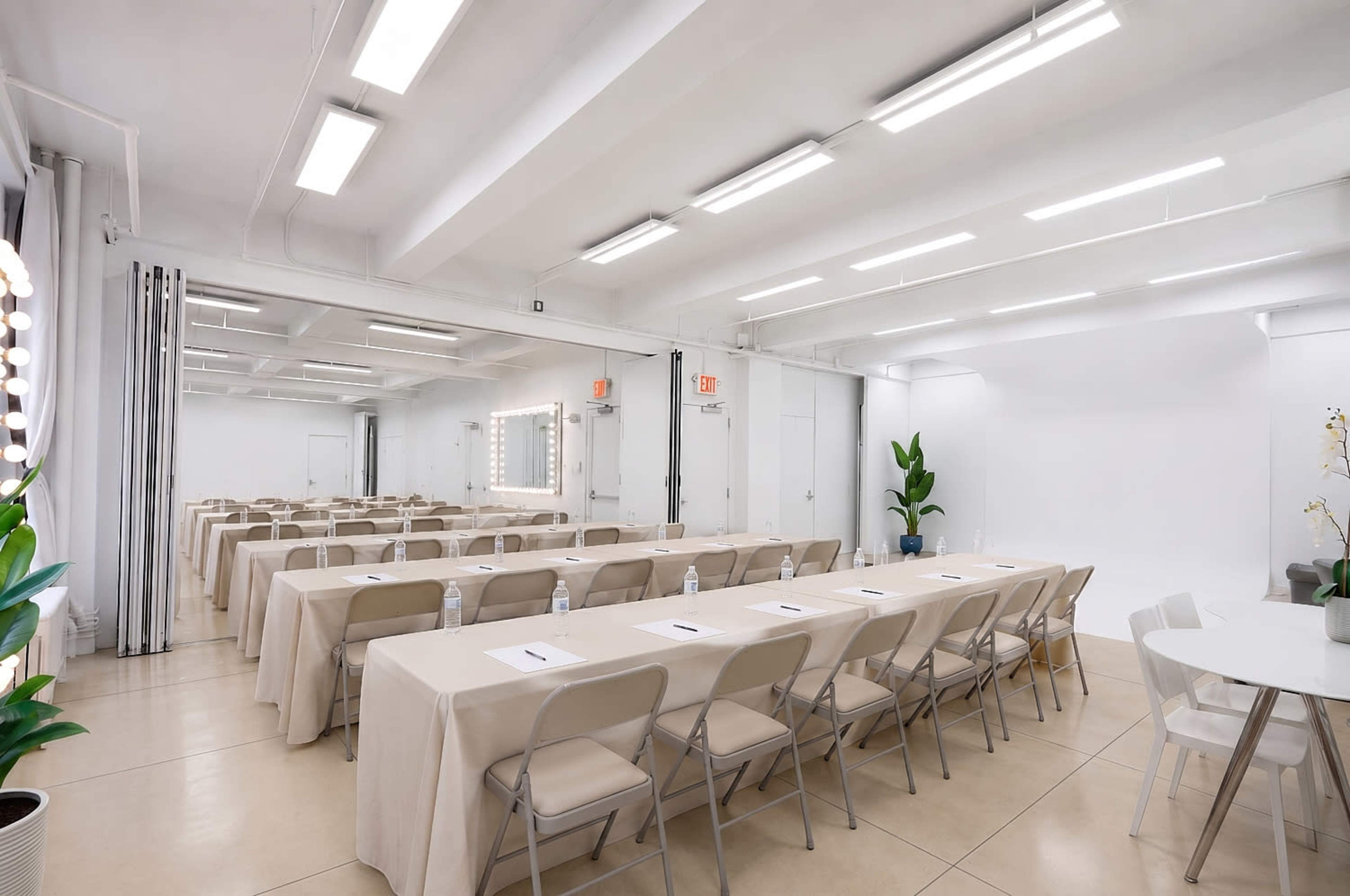 The image shows a spacious, well-lit meeting room with several rows of tables covered in beige tablecloths, facing a blank wall and large mirrors.