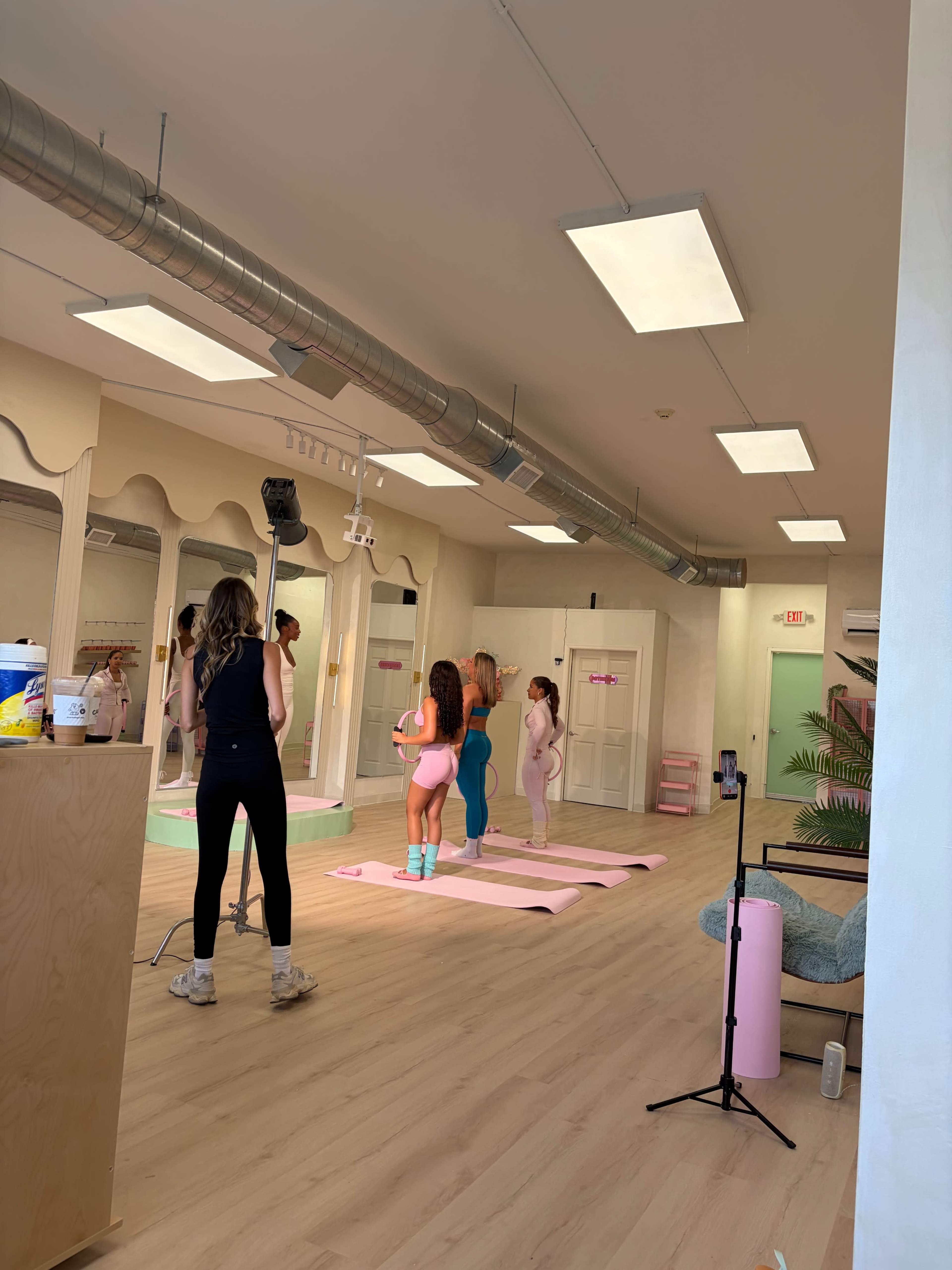 The image shows a brightly lit fitness studio with mirrored walls, where three individuals are engaged in a workout session while a photographer captures the scene.