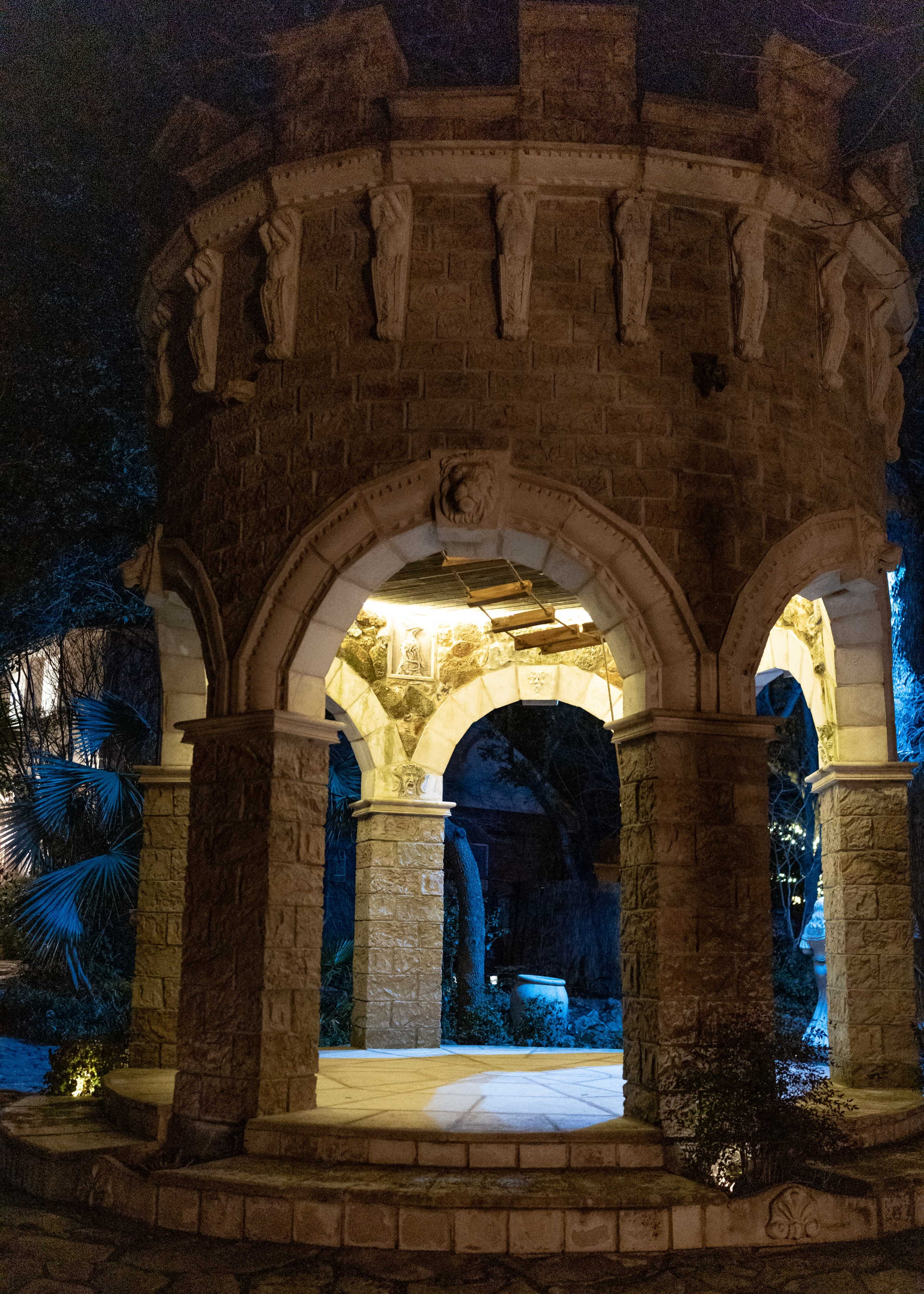 A stone gazebo with arched openings stands illuminated at night, featuring statuary figures around its upper section.