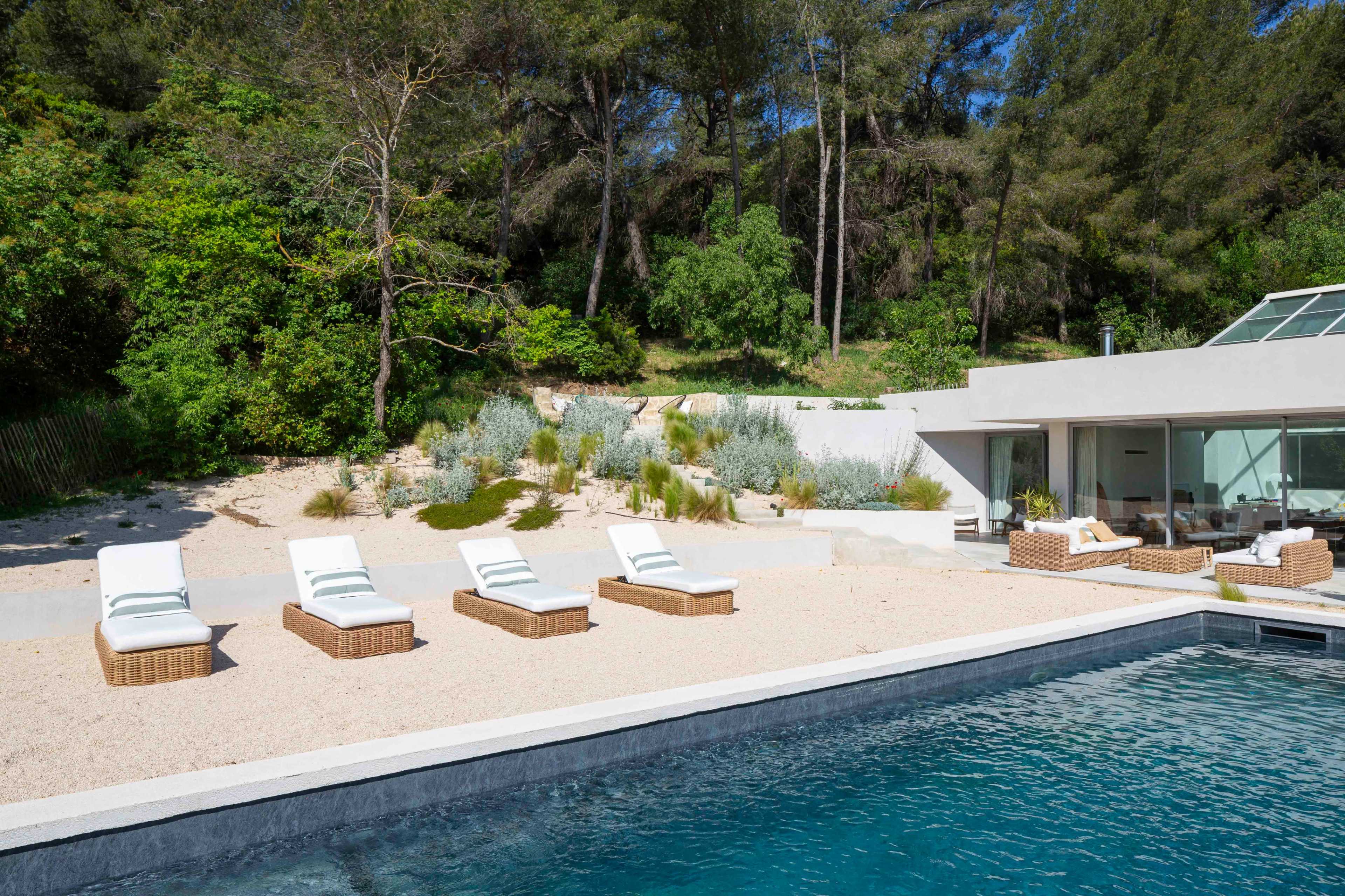 A modern poolside area features five white lounge chairs arranged on a sandy deck next to a clear blue pool, surrounded by greenery and a contemporary house.