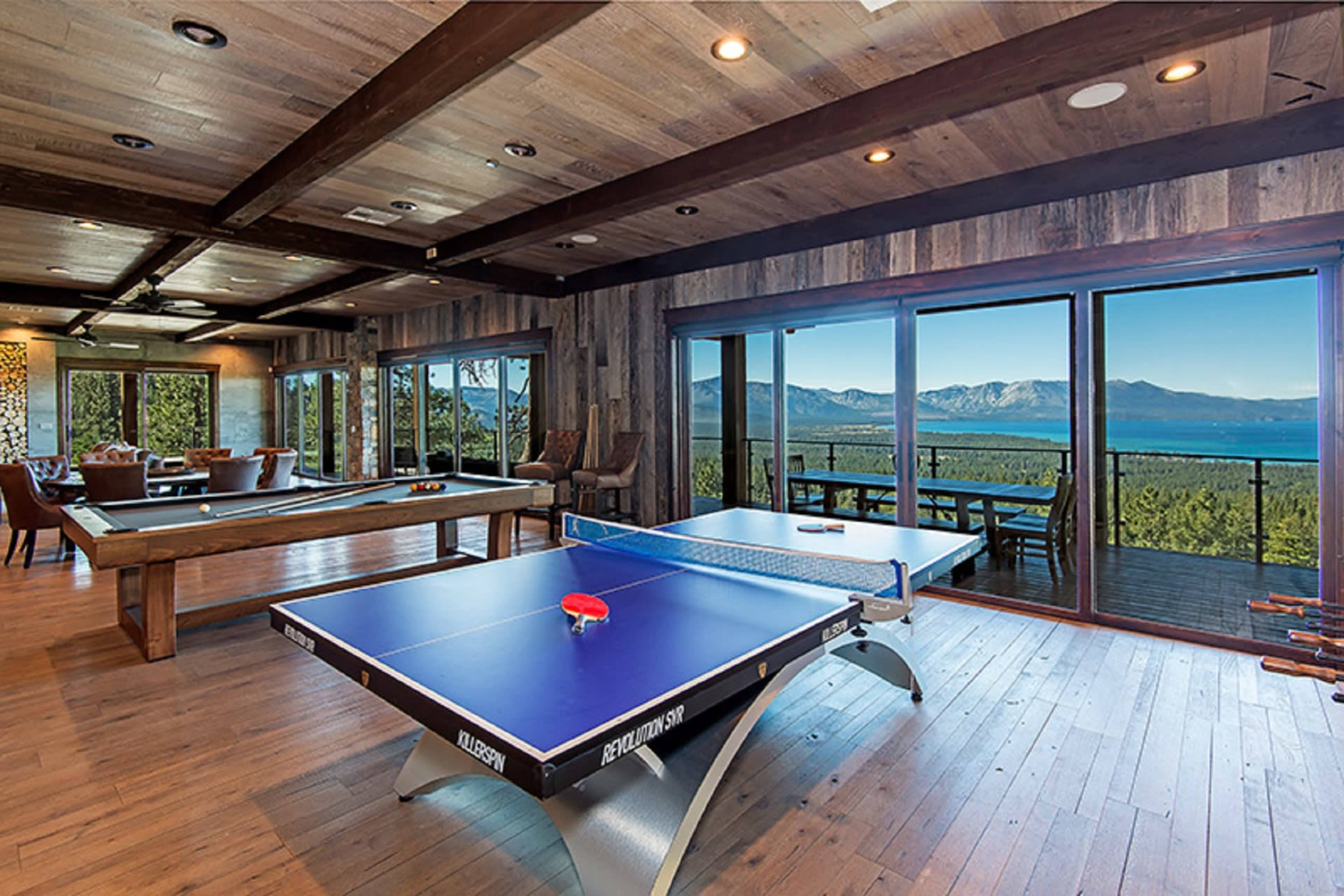 A game room featuring a ping pong table and a pool table, with large windows offering views of a lake and mountains in the background.