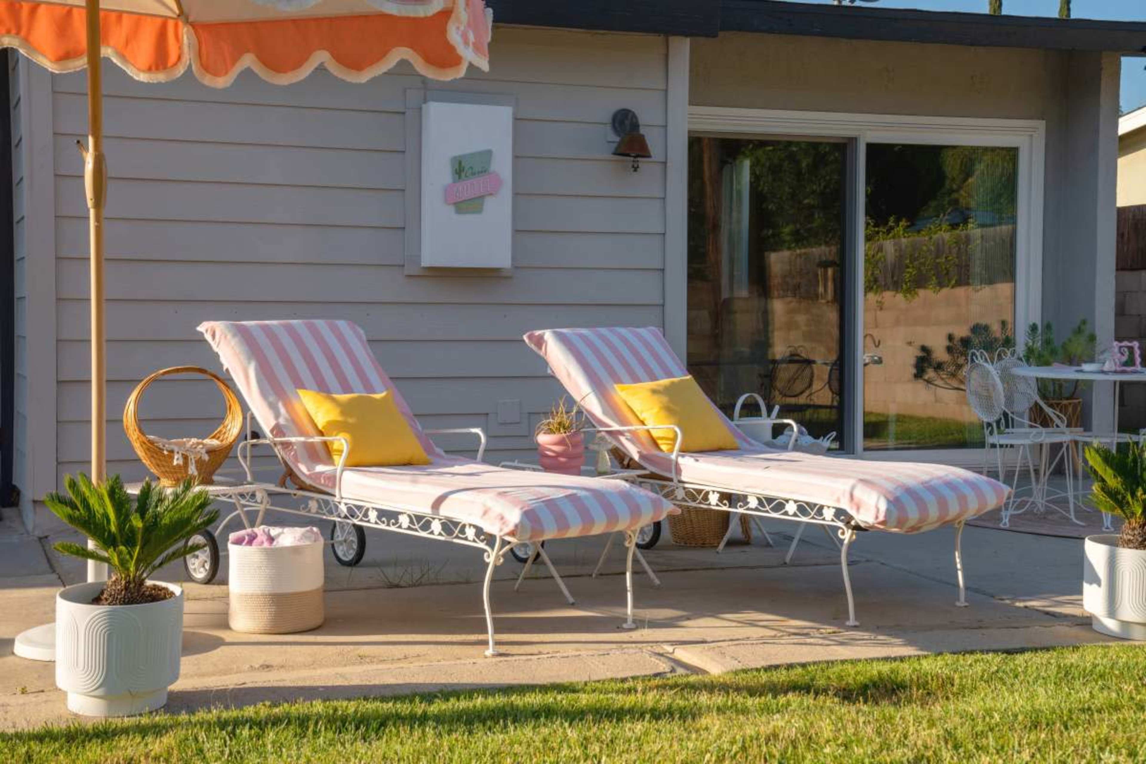 Two striped lounge chairs with yellow cushions are positioned beside a patio area featuring a small table and decorative plants under a sunny sky.