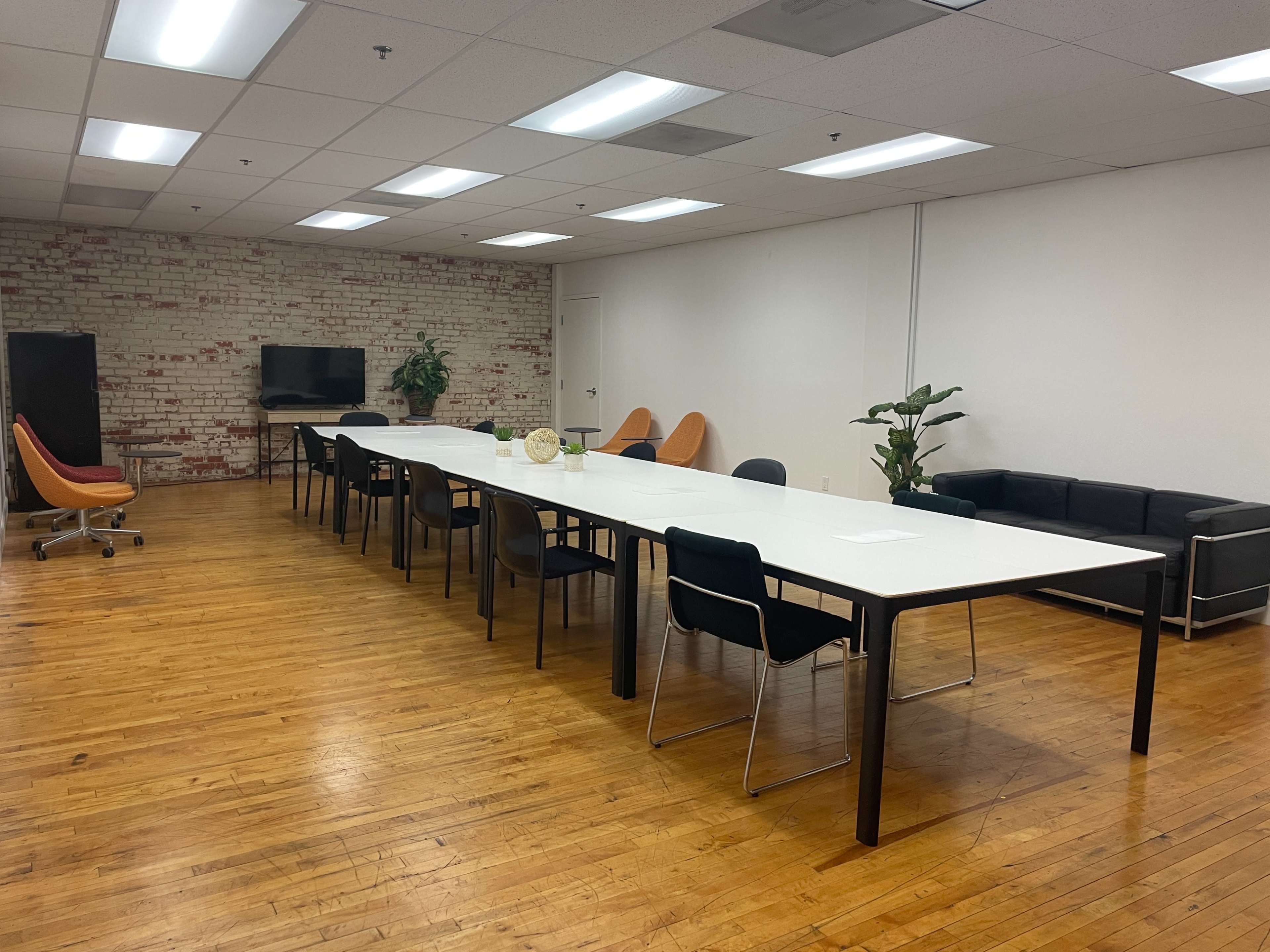 A spacious meeting room features a long white table surrounded by black and orange chairs, with a couch, television, and plants along the walls.