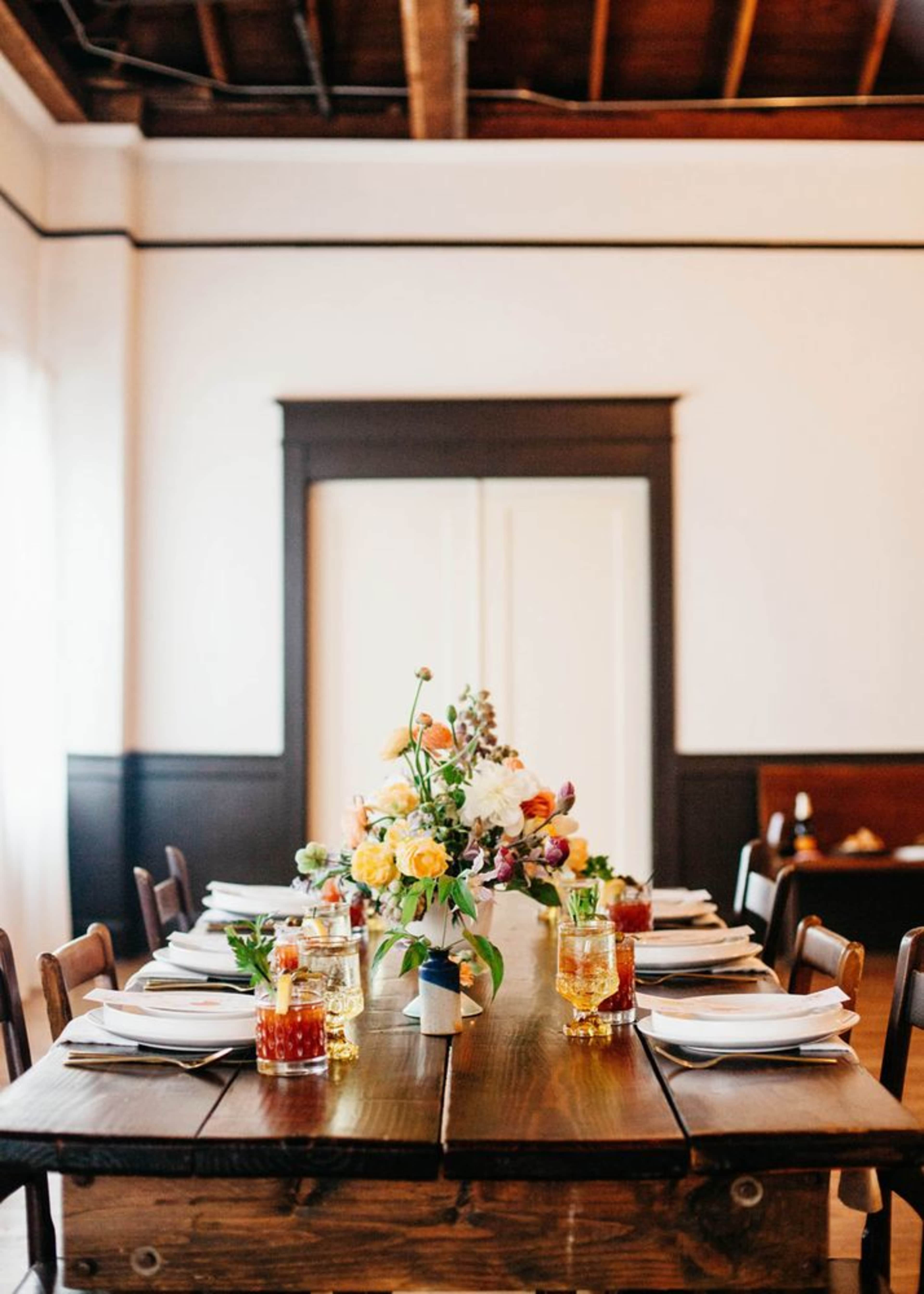 A long wooden table is set with plates and glasses, adorned with a vase of colorful flowers, in a well-lit dining room.