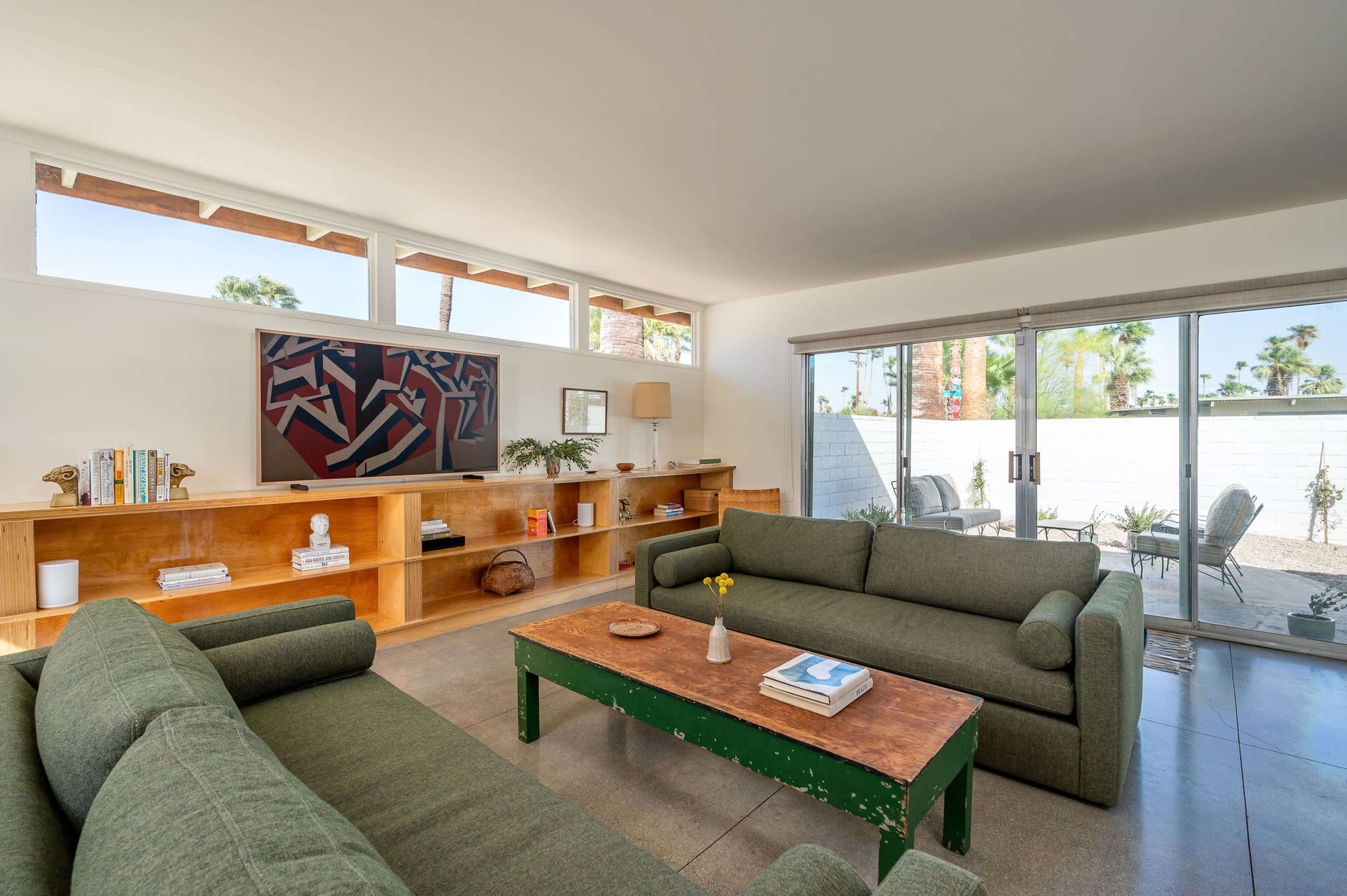 The image shows a modern living room with two green sofas facing a wooden coffee table, alongside a wall-mounted TV and a sliding glass door leading to an outdoor area.