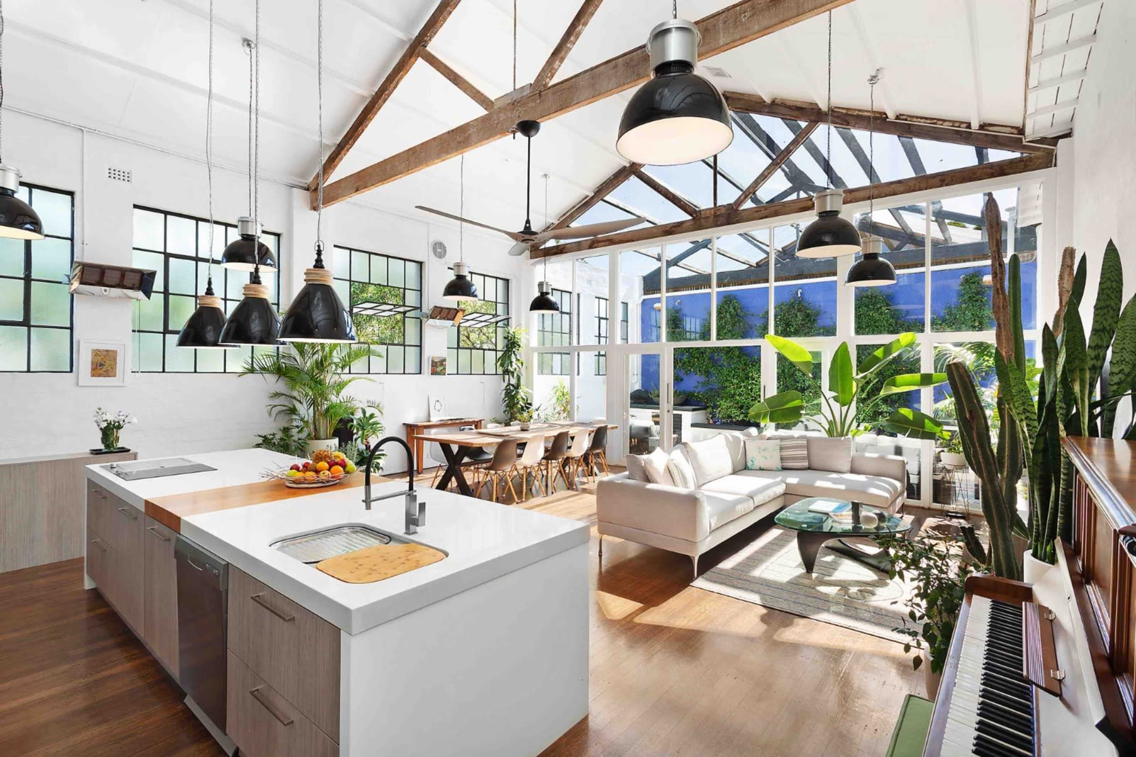 The image shows a modern kitchen and living area with large windows, wooden beams, plants, and a dining table.
