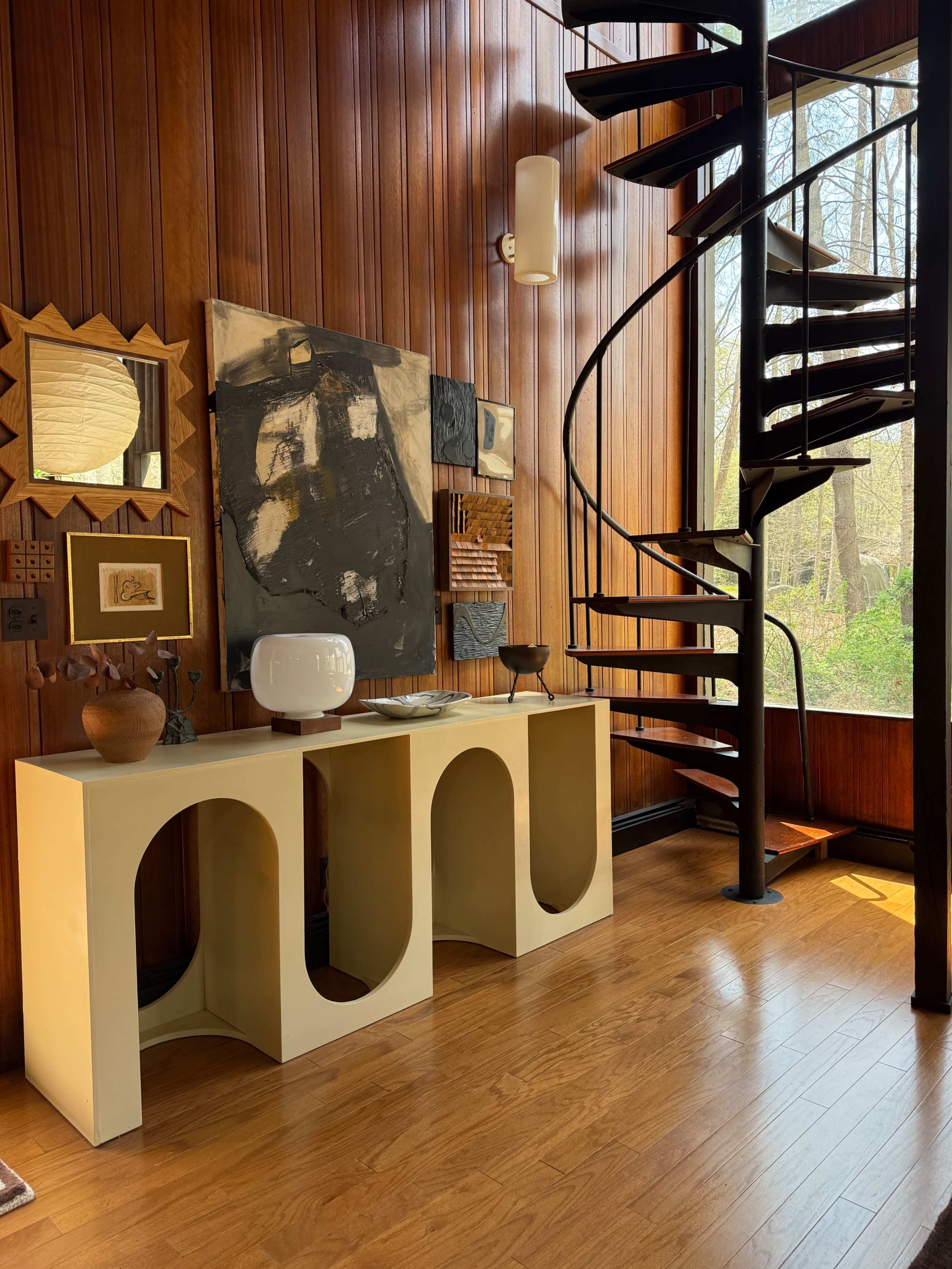 The image features a modern interior with a spiral staircase, wooden paneling, and various artworks displayed on the wall above a minimalist console table.