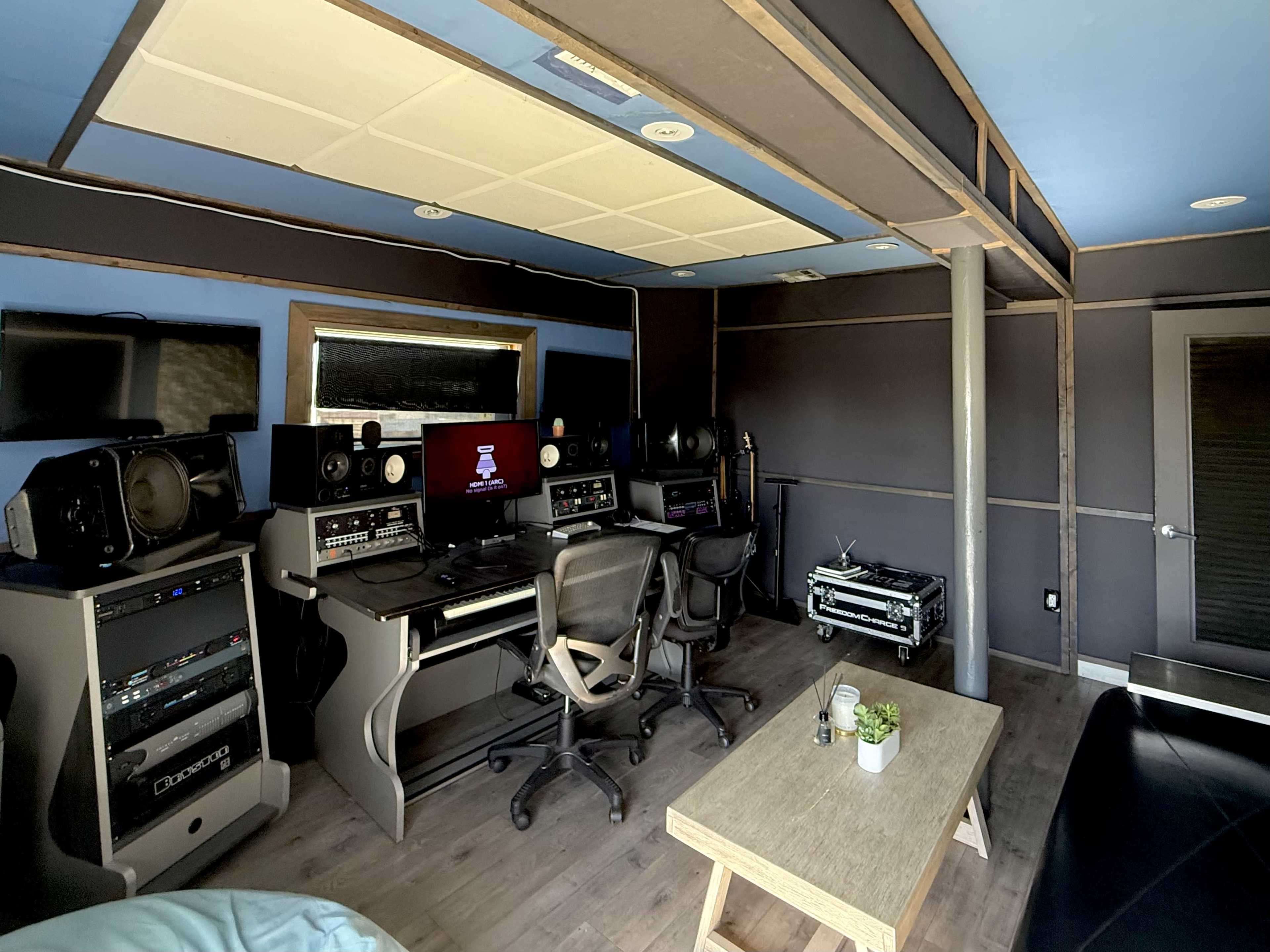 The image shows an audio recording studio equipped with mixing consoles, speakers, and a wooden table.
