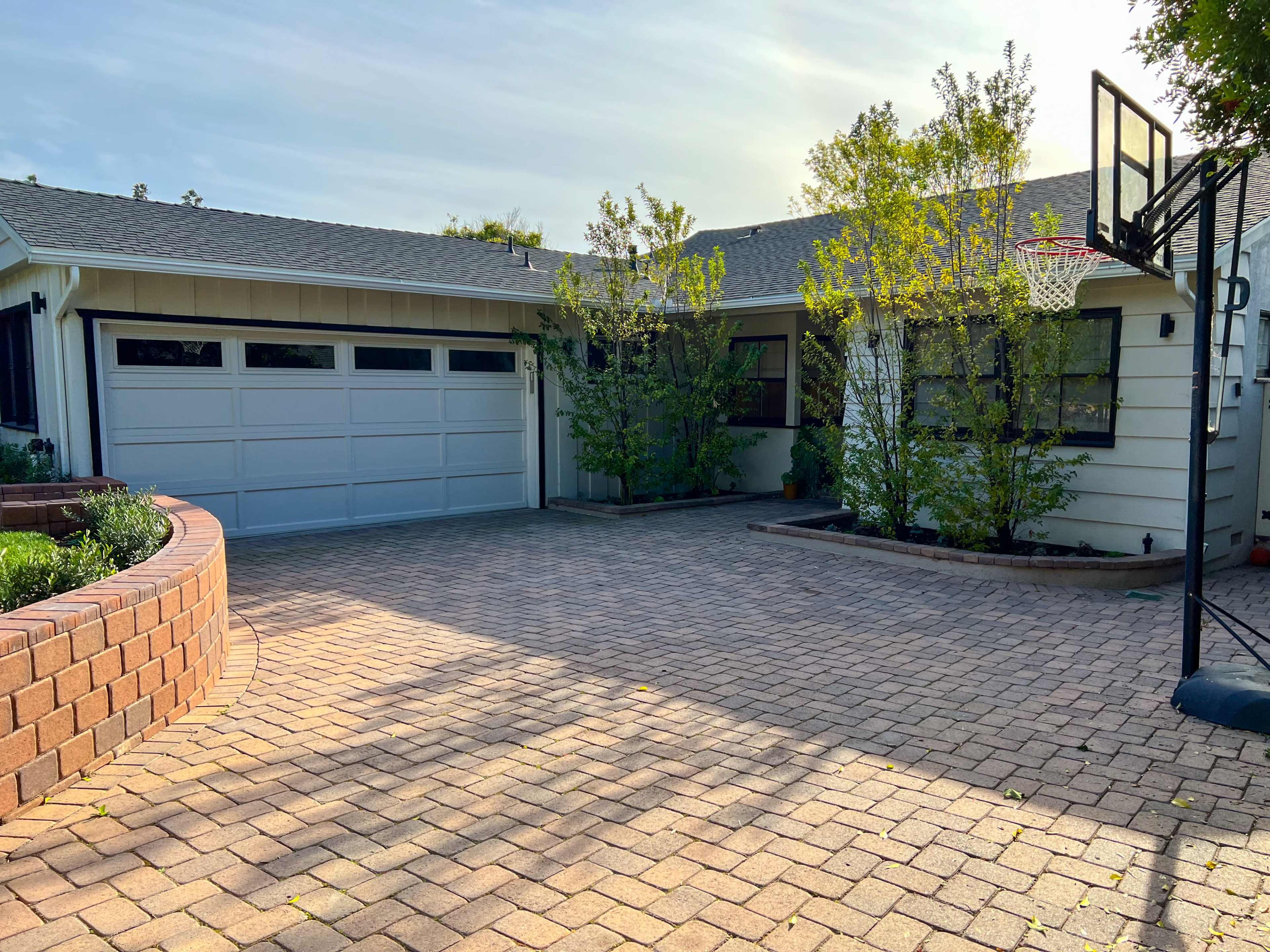 A house with a two-car garage, a driveway made of pavers, and a basketball hoop beside a landscaped area with small trees.