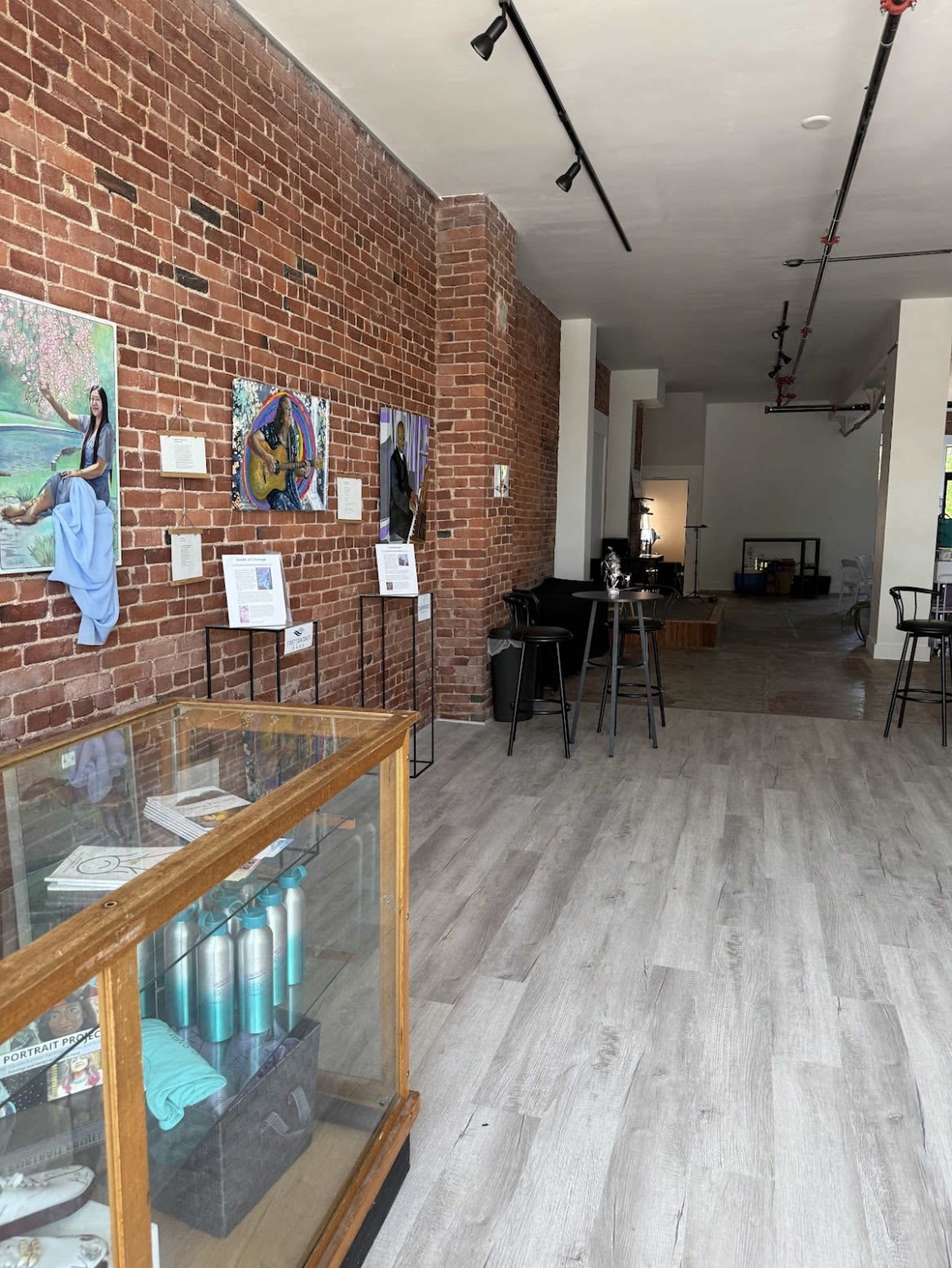 The image shows a modern gallery space with exposed brick walls, wood flooring, and various artworks displayed on easels.