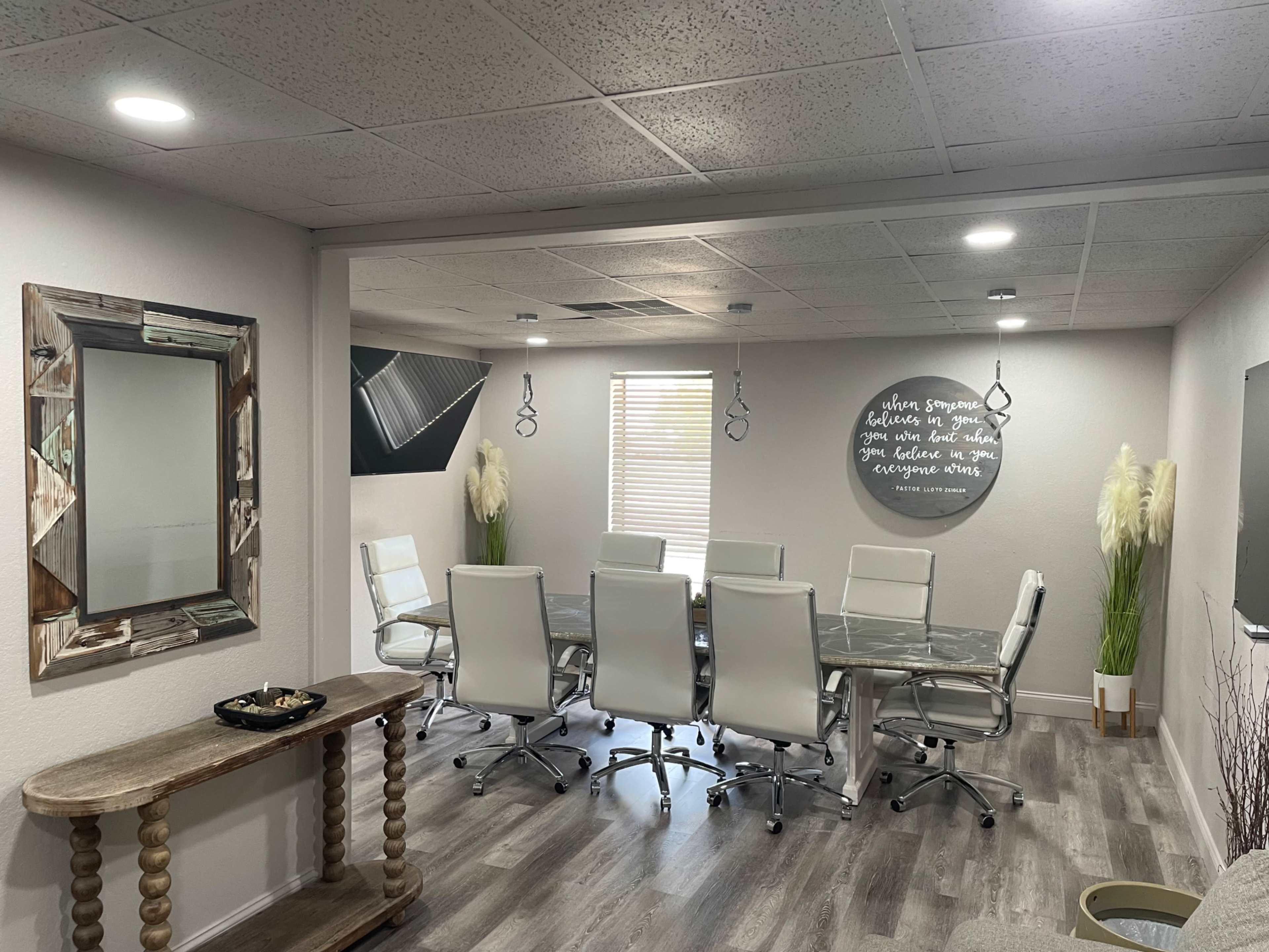 The image shows a modern conference room with a long glass table, white ergonomic chairs, and a wall adorned with decorative art and a motivational quote.