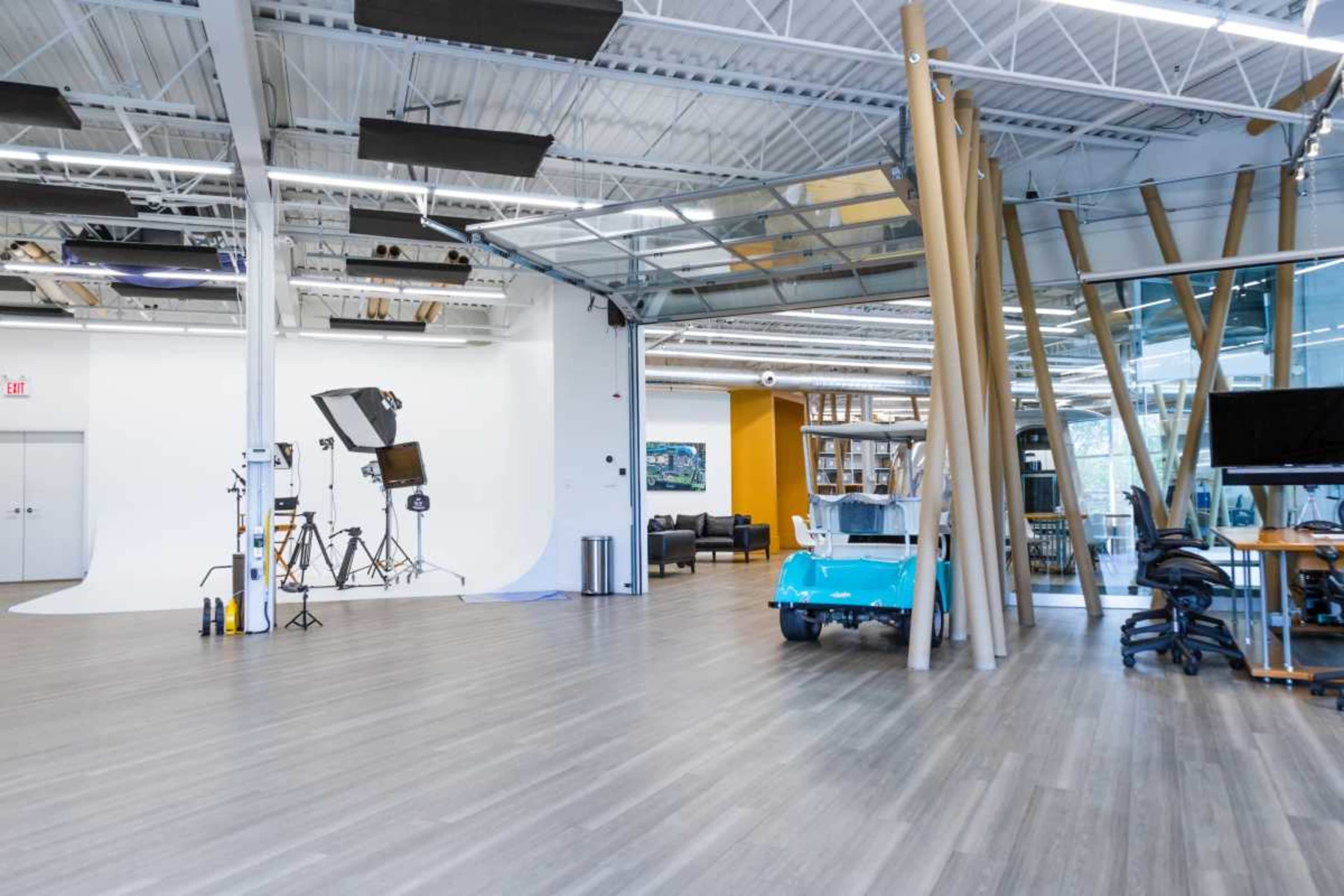 The image shows a modern, open studio space featuring a photography backdrop, lighting equipment, and a colorful cart amidst a mix of seating areas and workstations.