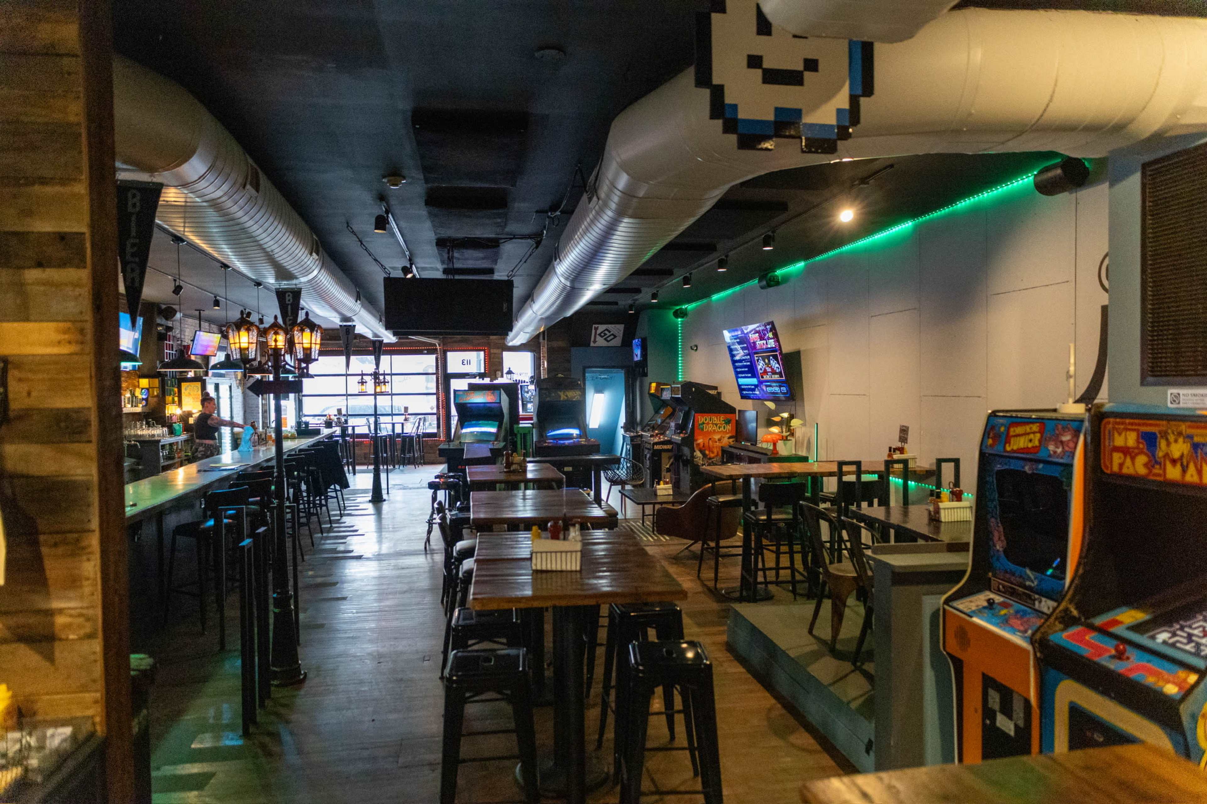 The image shows a spacious bar and arcade with rows of tables, arcade machines, and illuminated wall sections.
