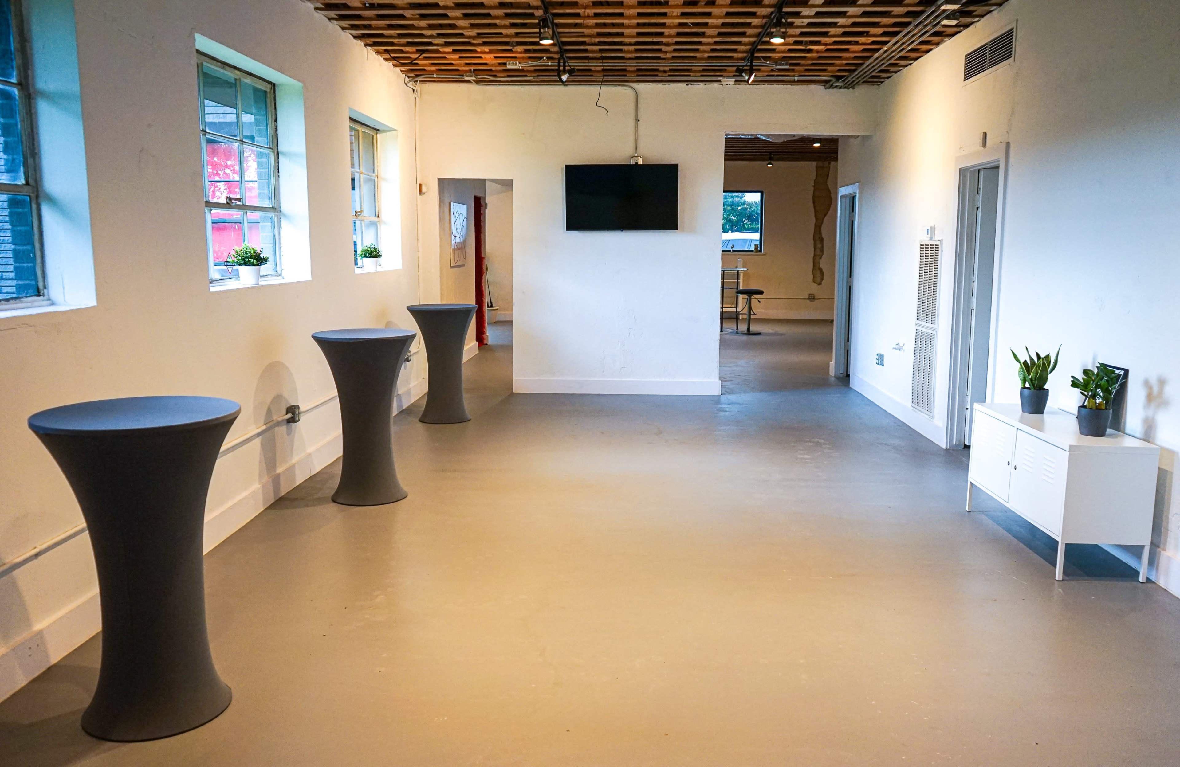 The image shows a spacious, minimally decorated room with three tall tables positioned along one wall and a flat-screen television mounted on another wall.