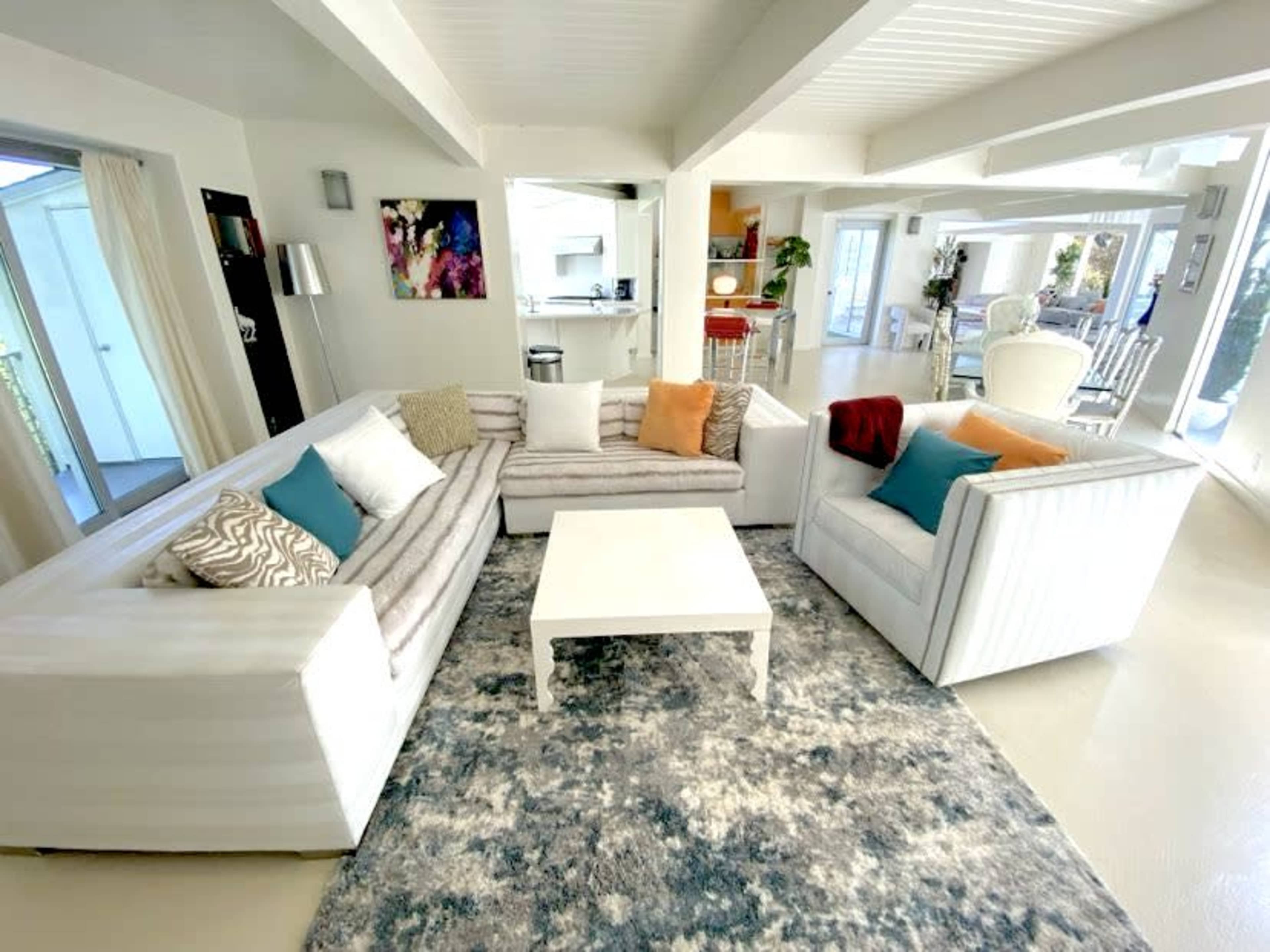 The image shows a modern living room with white sectional sofas, an area rug, and a coffee table, all set in a light-filled space that opens to a kitchen area in the background.