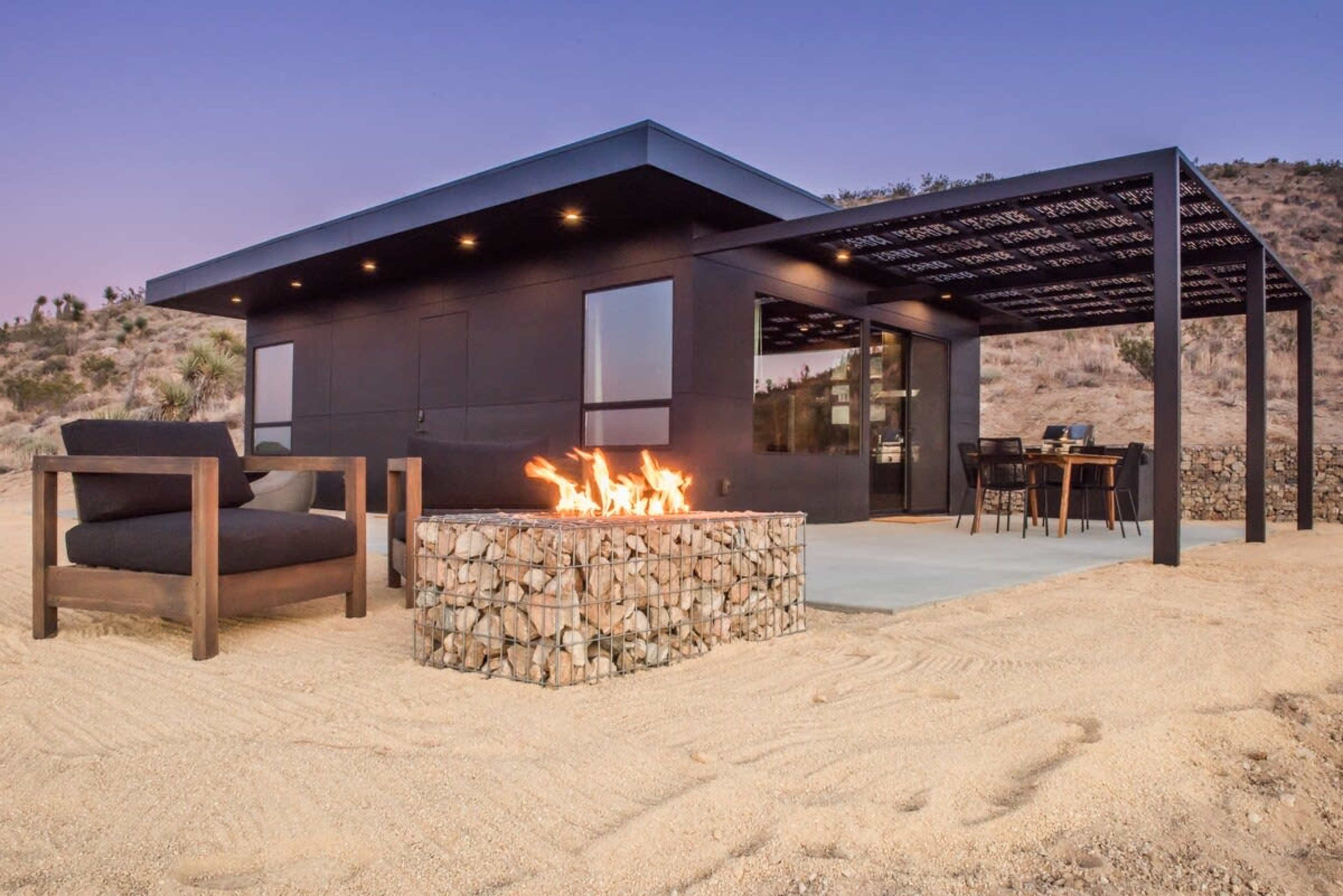 A modern black building with large windows and a covered patio is situated in a sandy desert landscape, featuring a stone fire pit in front.