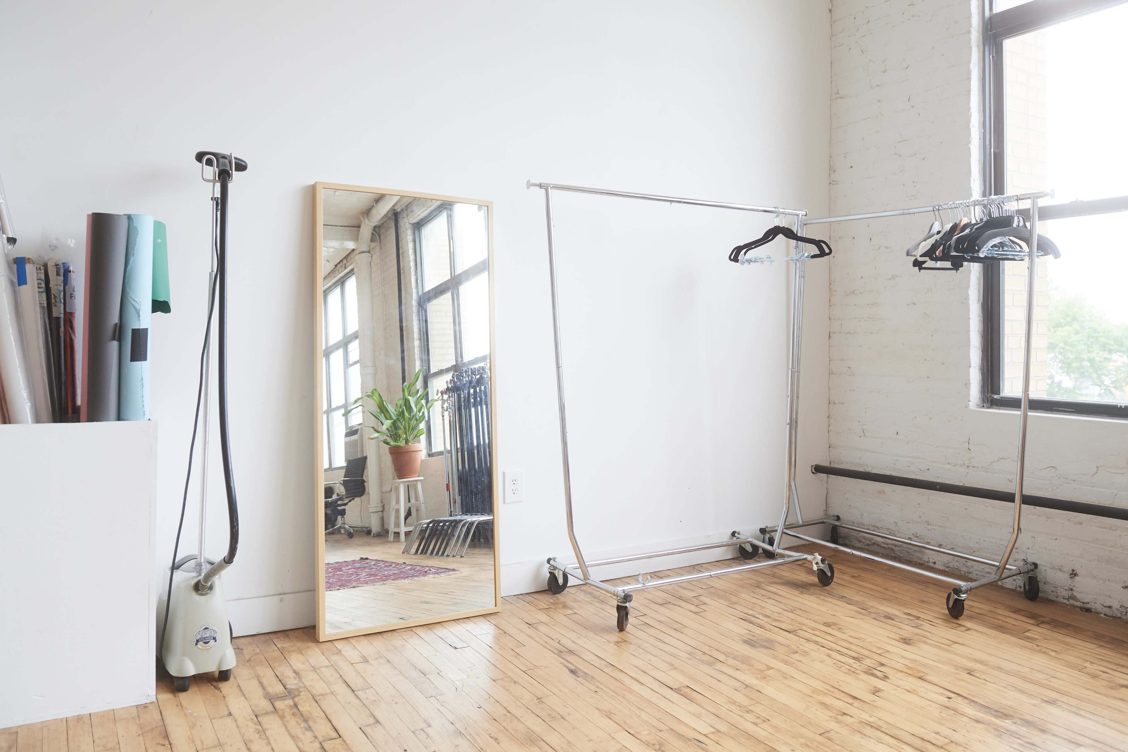 A bright room with a large mirror, a clothing rack on wheels, and a garment steamer beside a wall of windows.