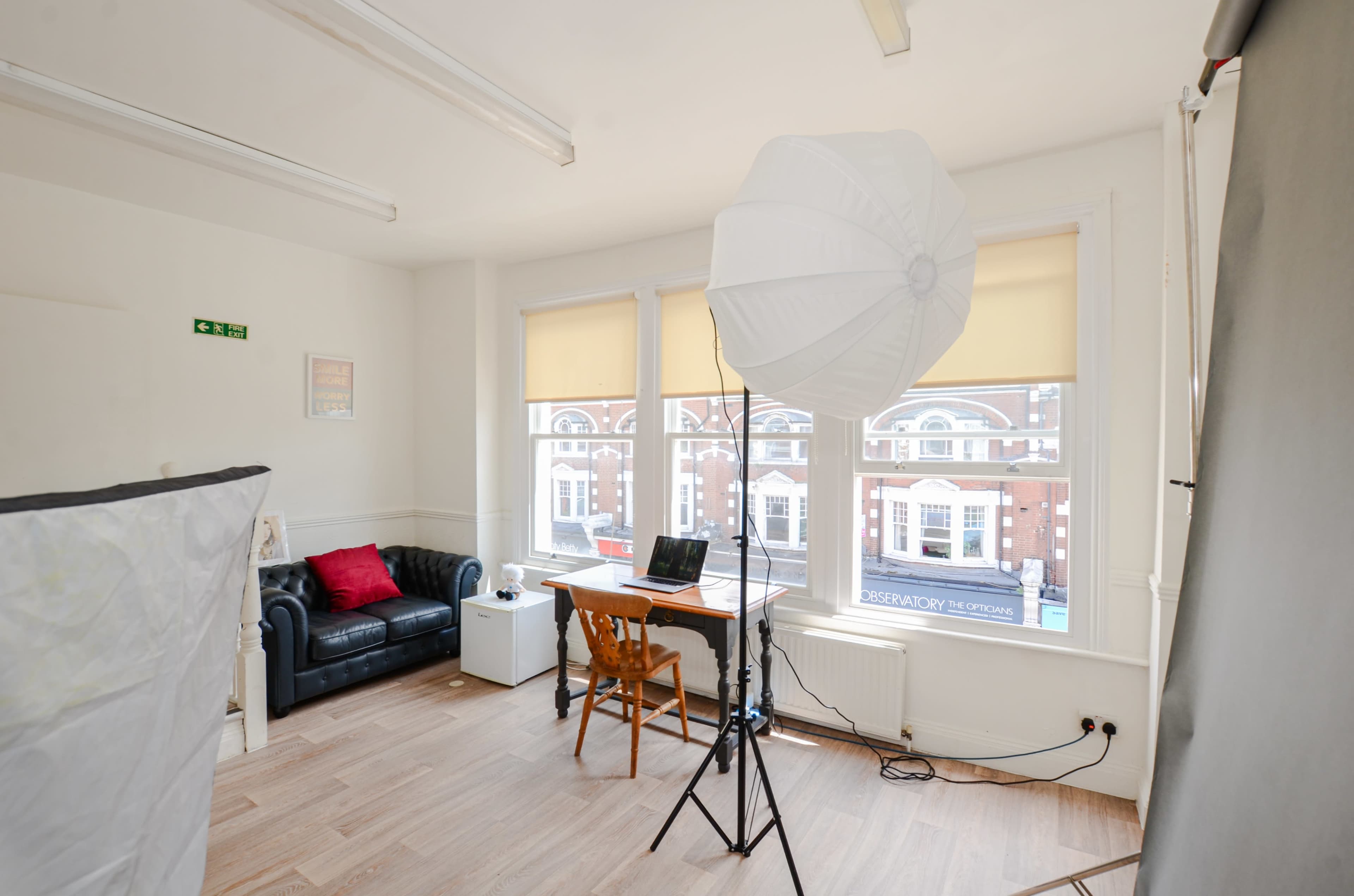 The image shows a well-lit interior of a studio or office space featuring a black leather sofa, a wooden desk with a laptop, and a large softbox light.