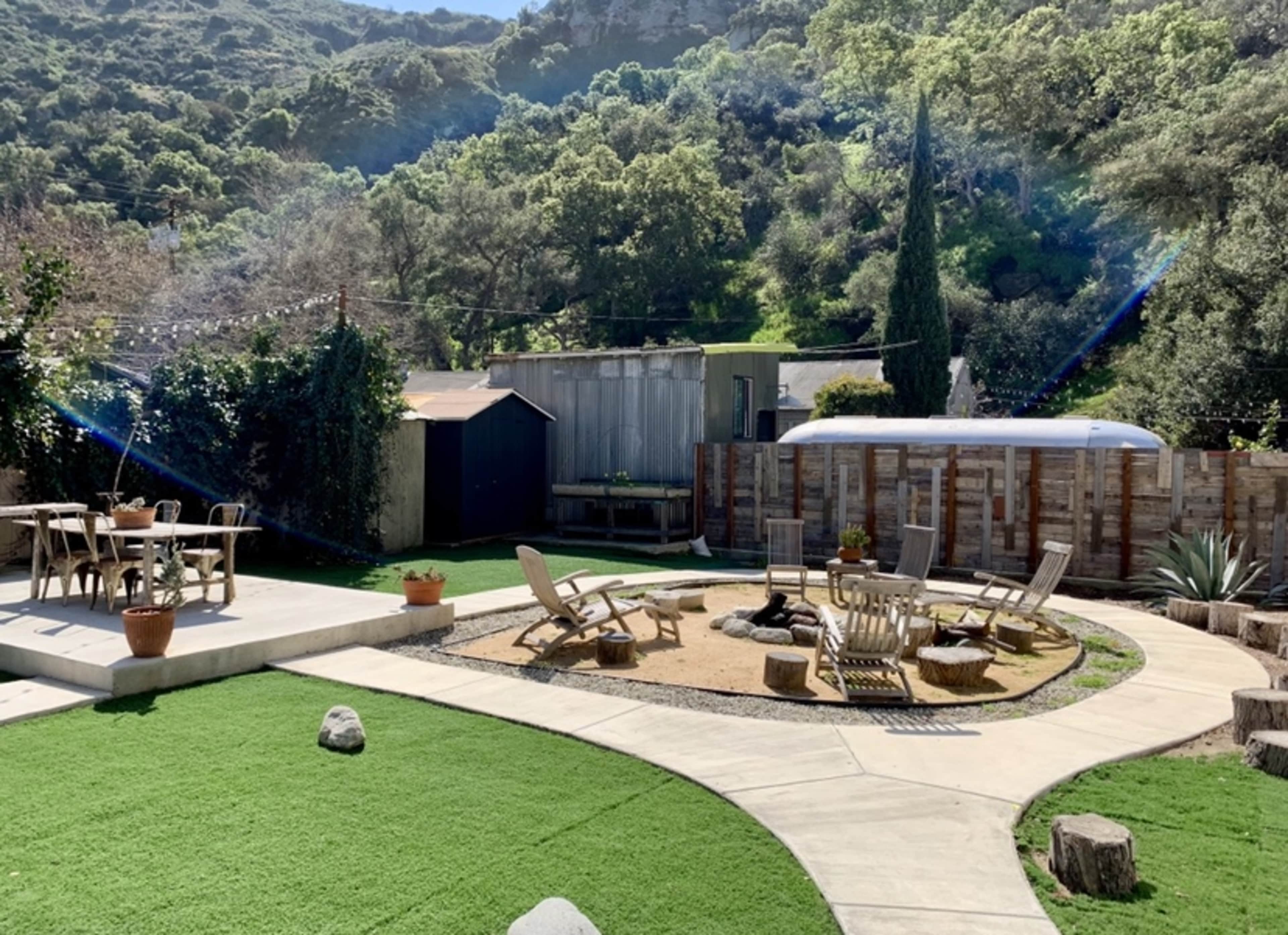 A landscaped outdoor area featuring seating around a fire pit, a grassy lawn, and a shed in a natural setting with hills in the background.
