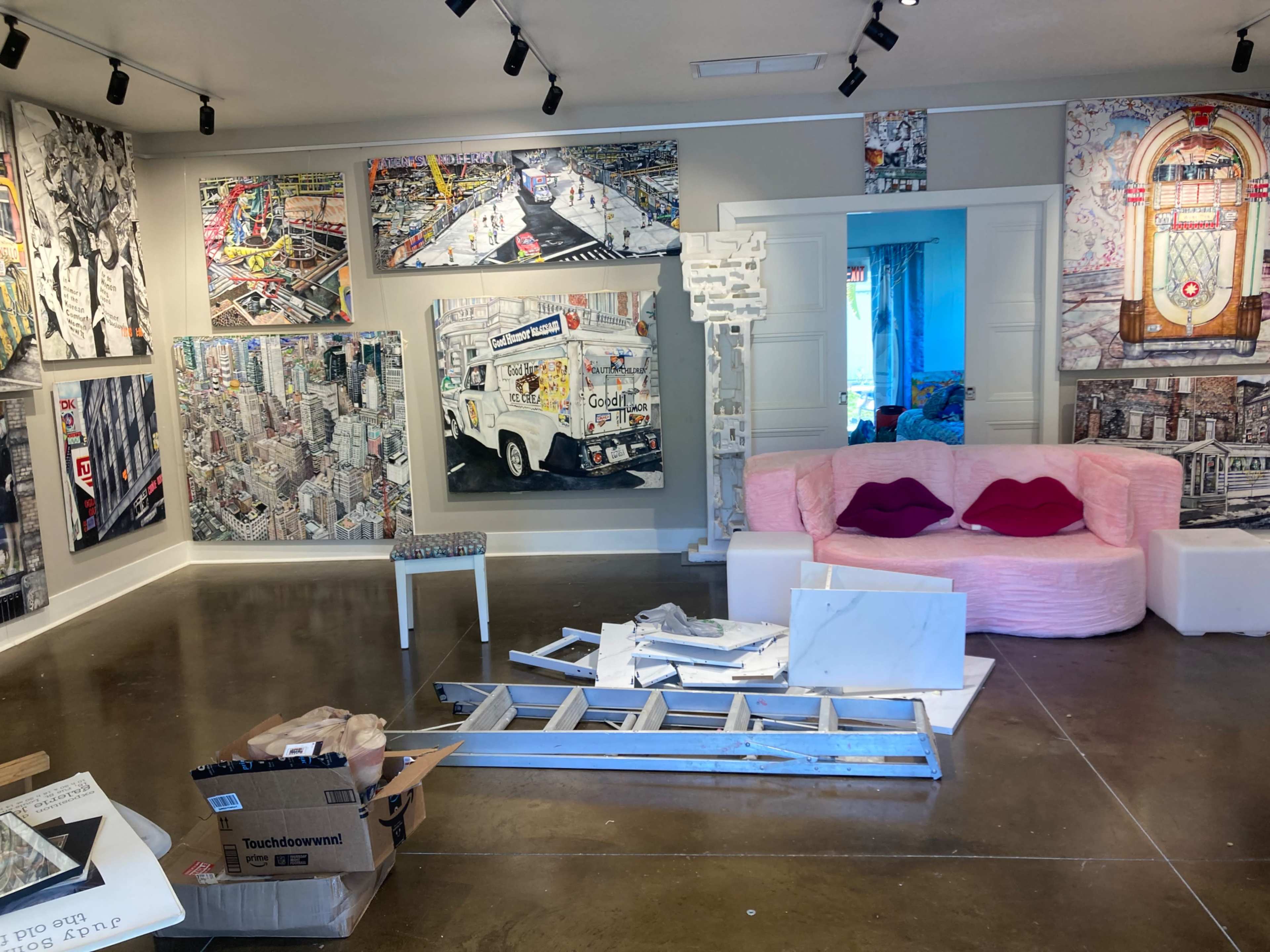The image depicts an art studio featuring various large urban-themed artworks on the walls, a pink couch with heart-shaped pillows, and scattered tools and materials around the floor.