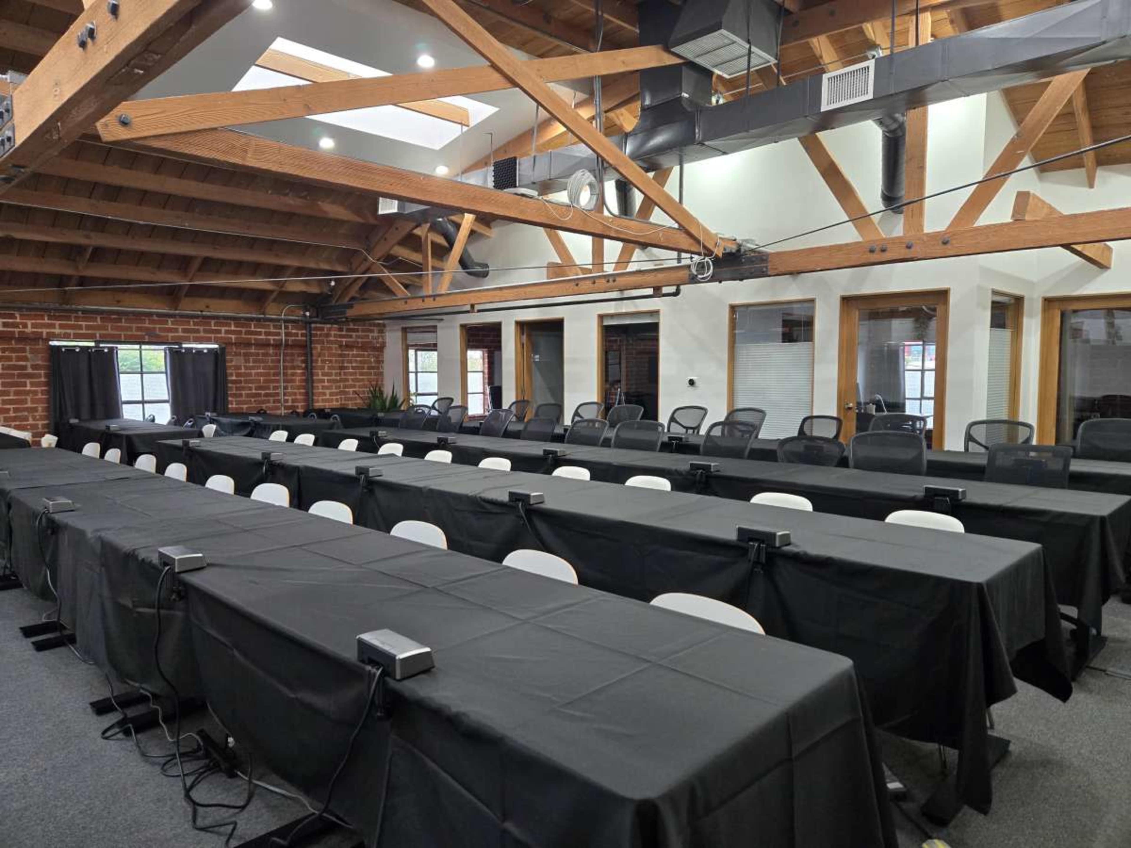 The image shows a spacious meeting room featuring rows of long black tables with chairs on either side, illuminated by natural light from overhead windows.