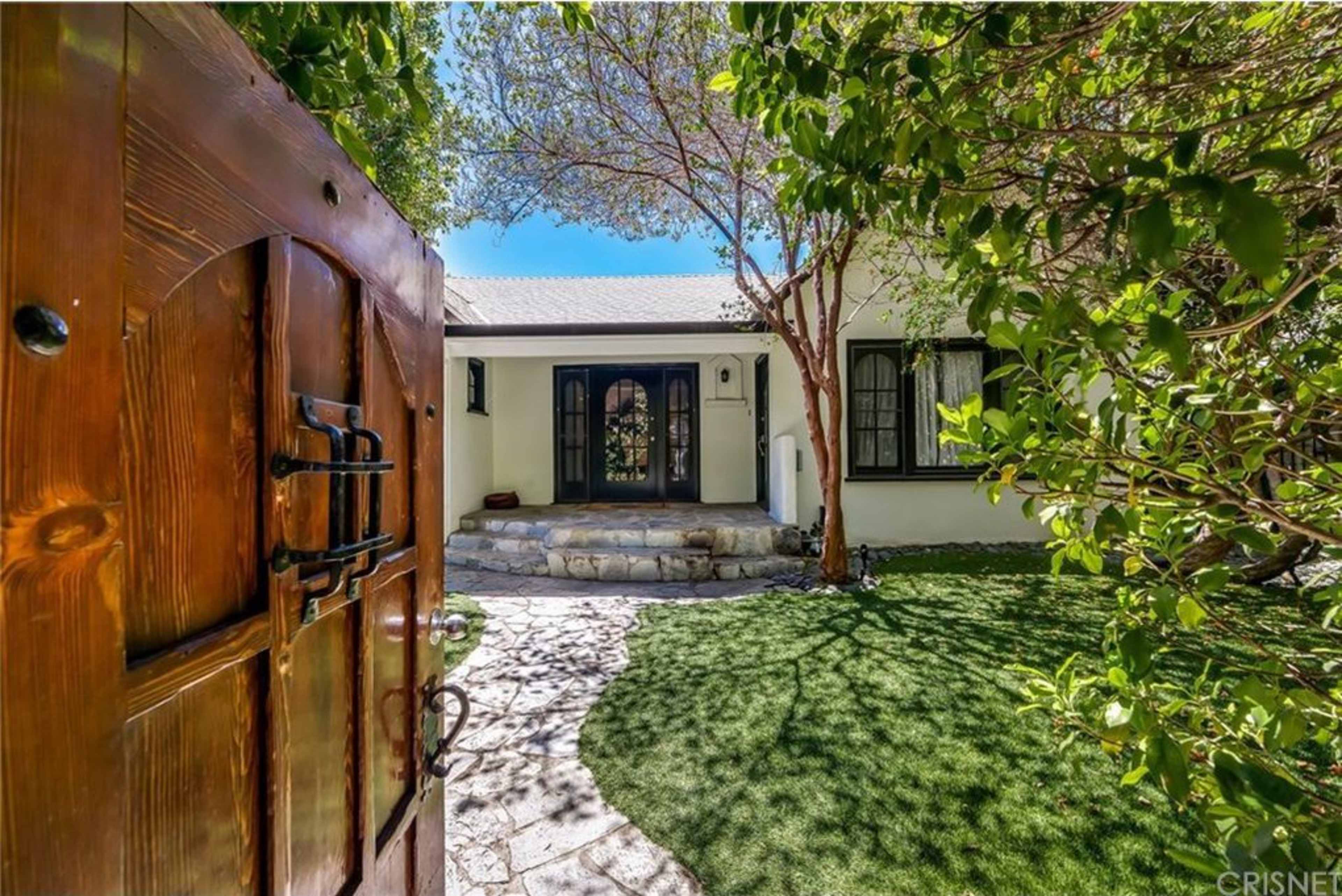 The image shows a stone pathway leading to a house with a wooden front door, surrounded by green foliage.