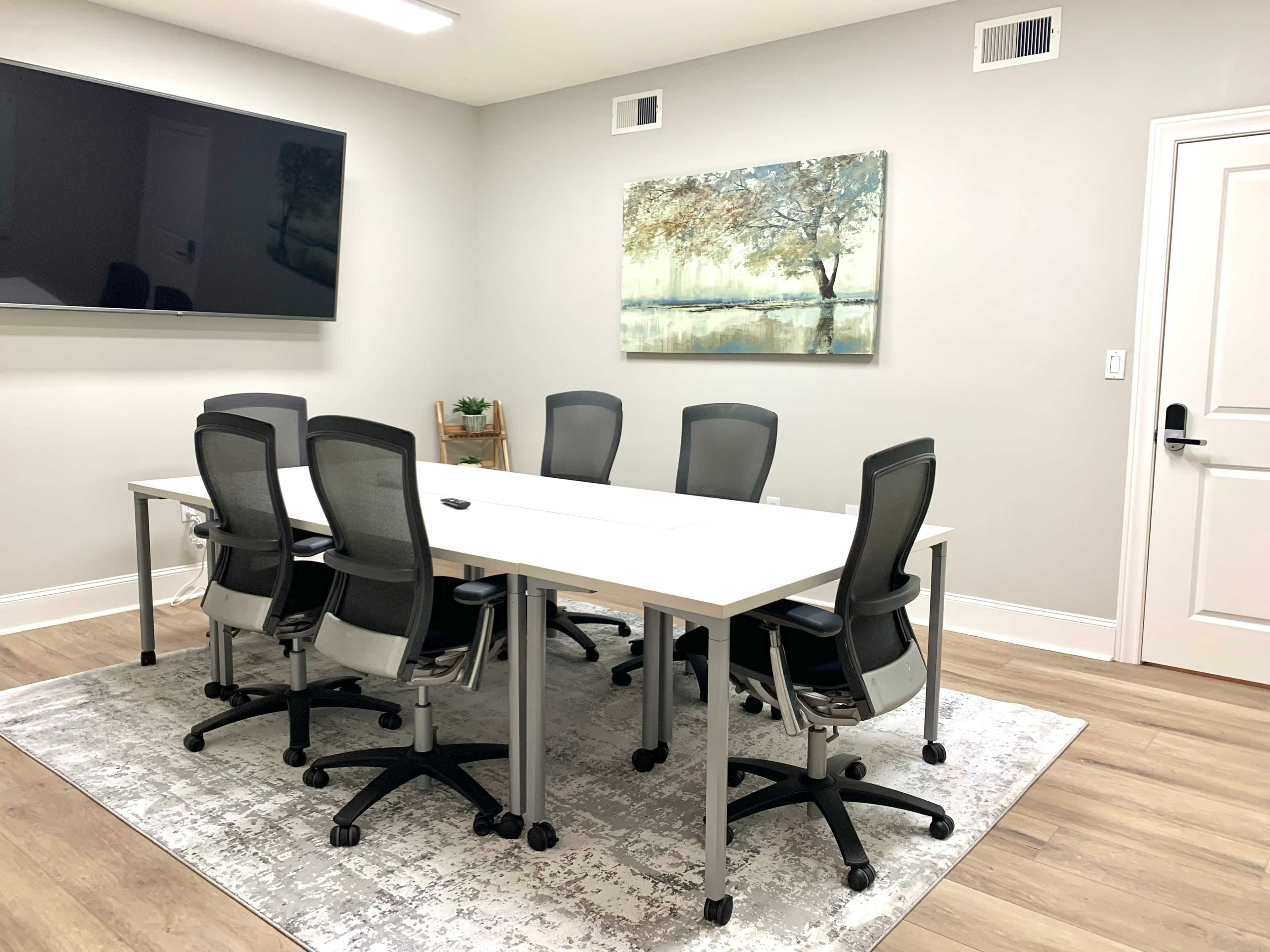 A modern conference room with a large table surrounded by six ergonomic chairs, a wall-mounted TV, and an artwork depicting a tree.