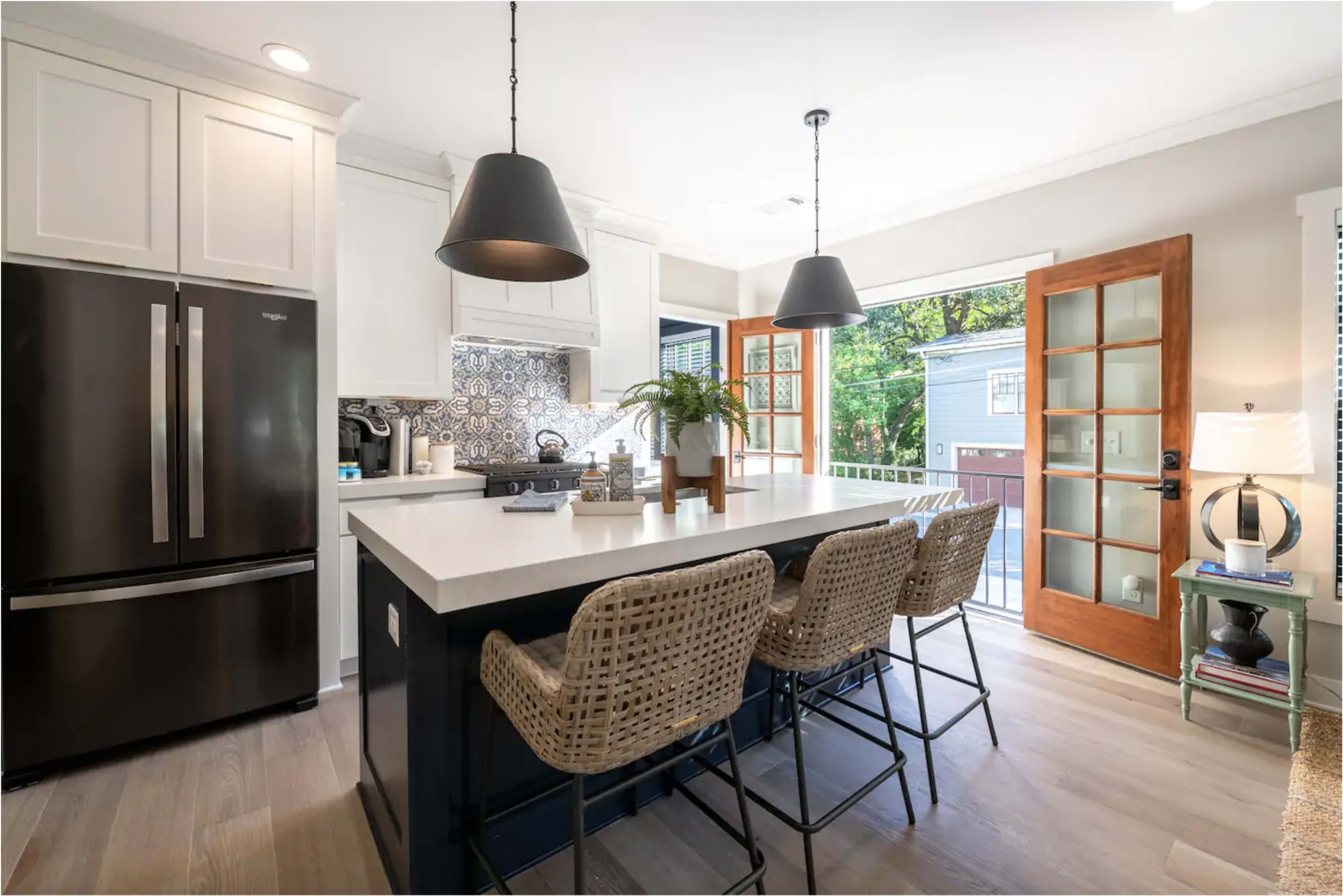 The image shows a modern kitchen with a large island featuring bar stools, black appliances, and double French doors opening to an outdoor space.
