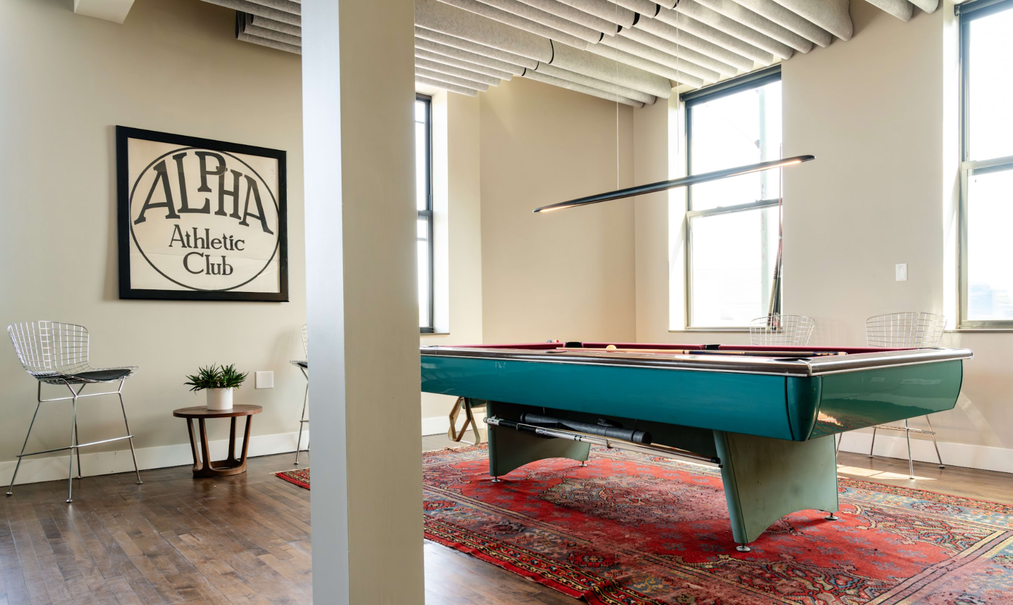 The image features a pool table in a well-lit room with large windows, surrounded by two wire chairs and a decorative rug, displaying a framed sign that reads "ALPHA Athletic Club."