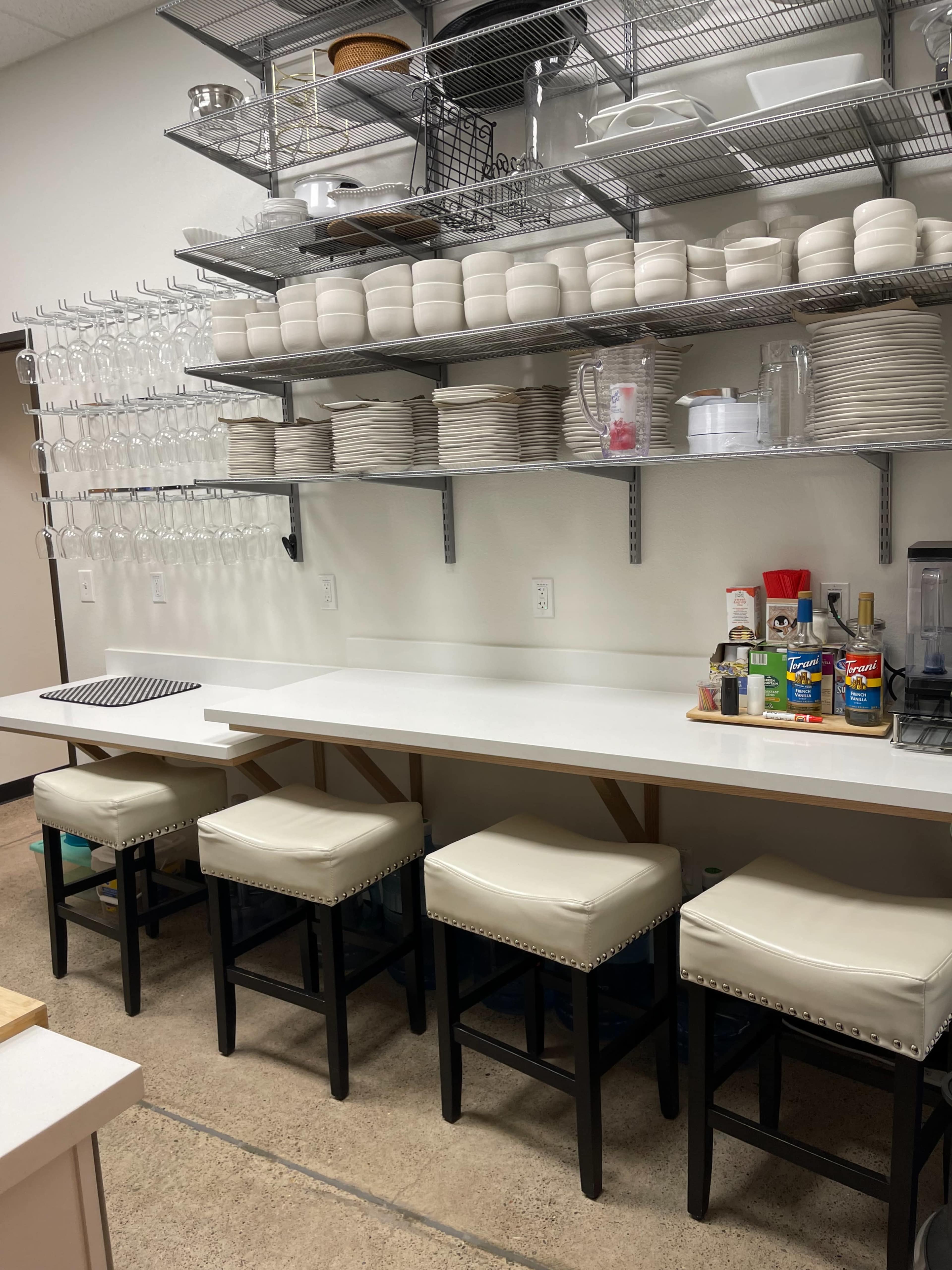 A clean kitchen space features a wall of shelves stocked with stacked dishes and glassware, complemented by a white countertop and four barstools.
