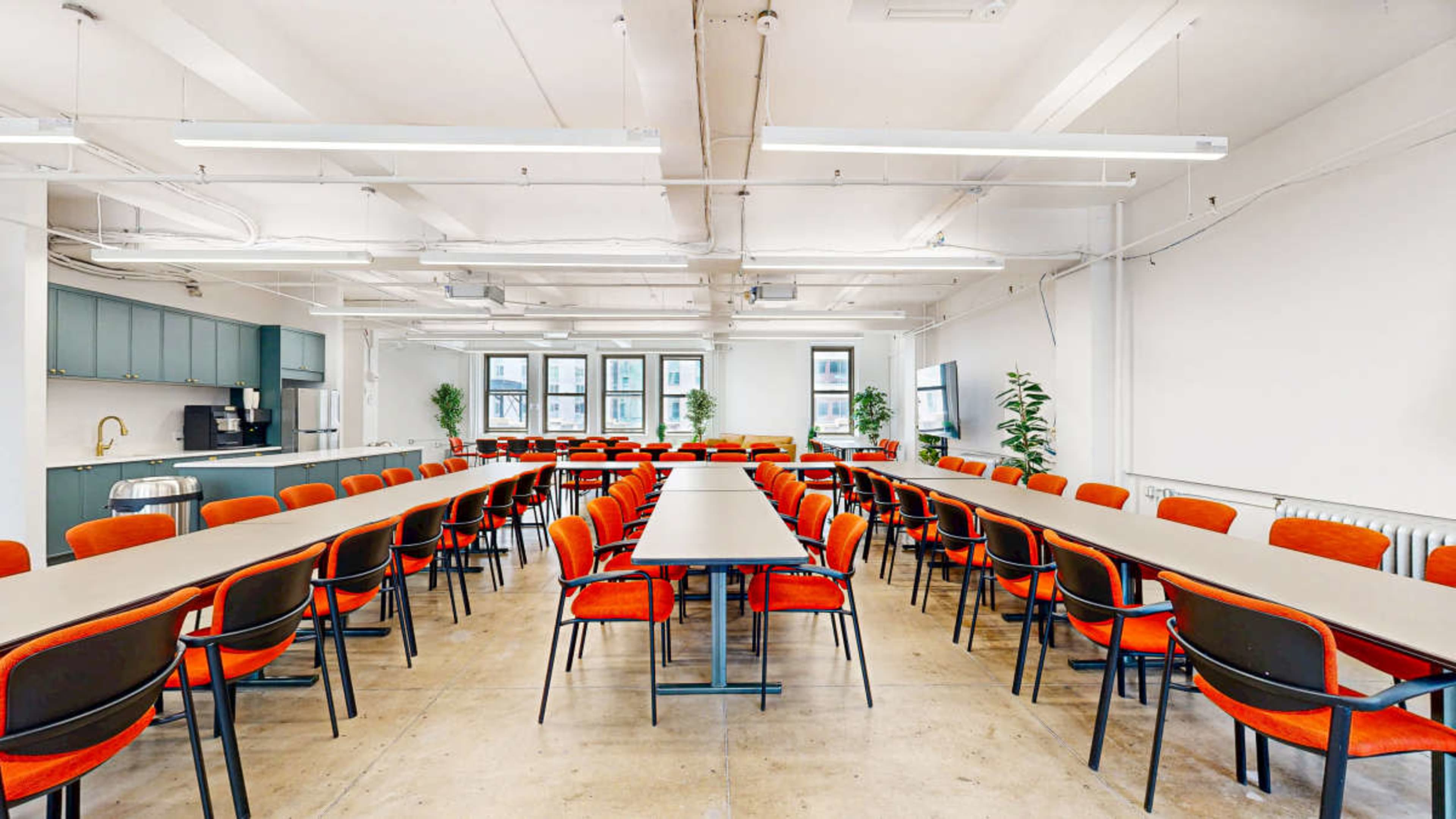 The image shows a spacious conference room with long tables arranged in rows and orange chairs, complemented by a kitchenette in one corner.