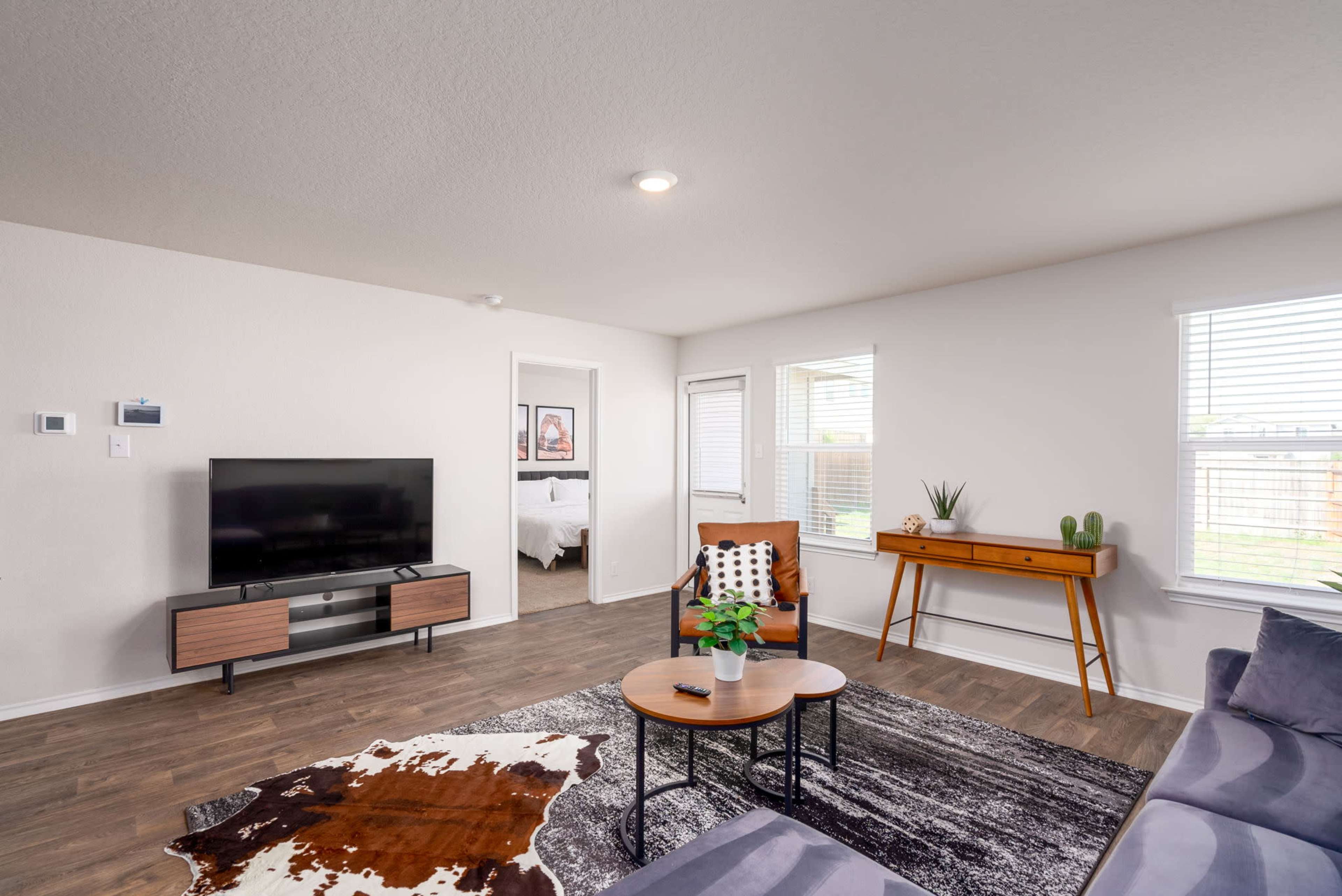 A modern living room features a television on a wooden stand, a grey sofa, two coffee tables, and large windows with natural light.
