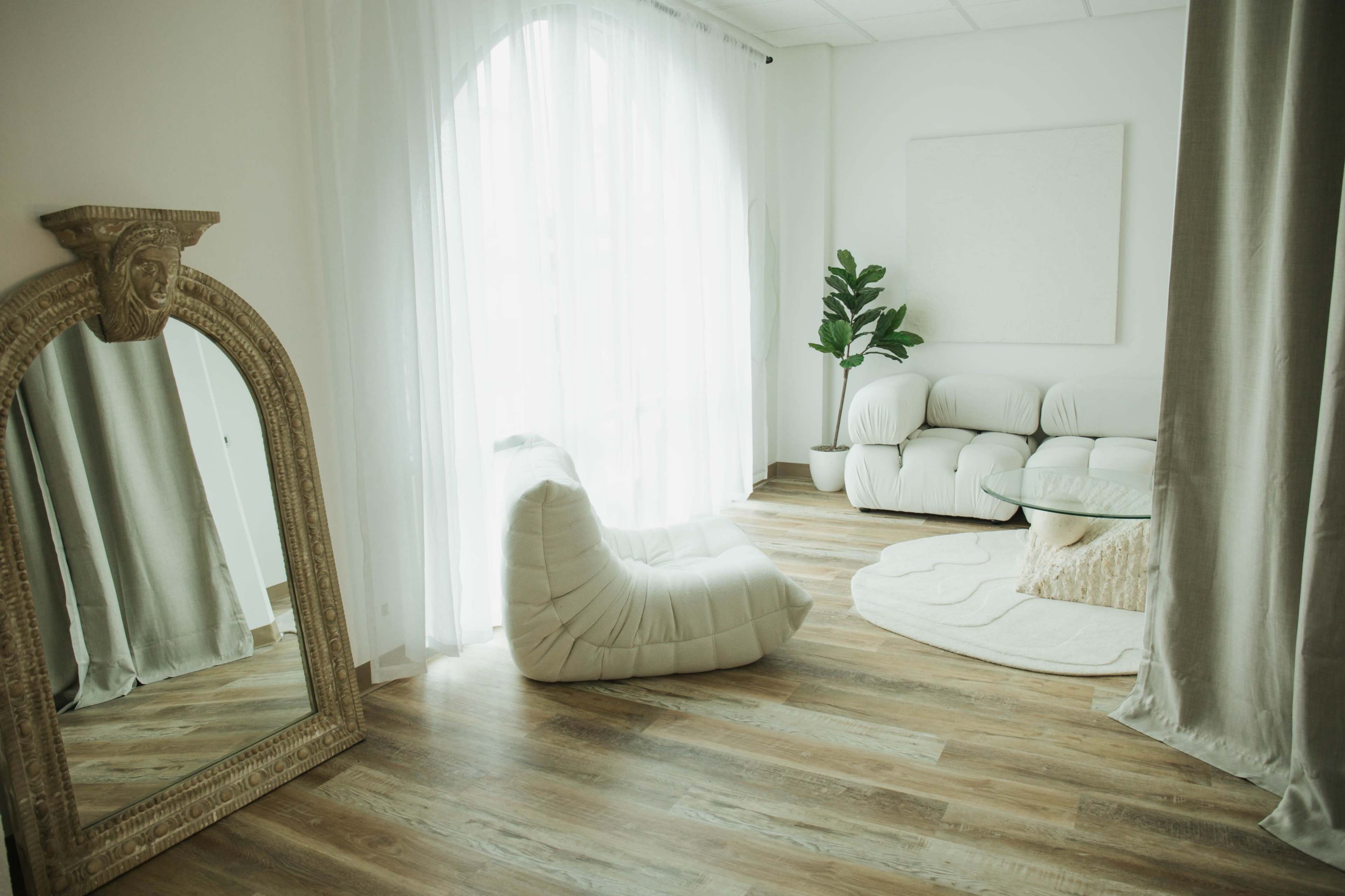 The room features a large mirror, a plush seating area with a chair and sofa, and a soft rug, all illuminated by natural light filtering through sheer curtains.