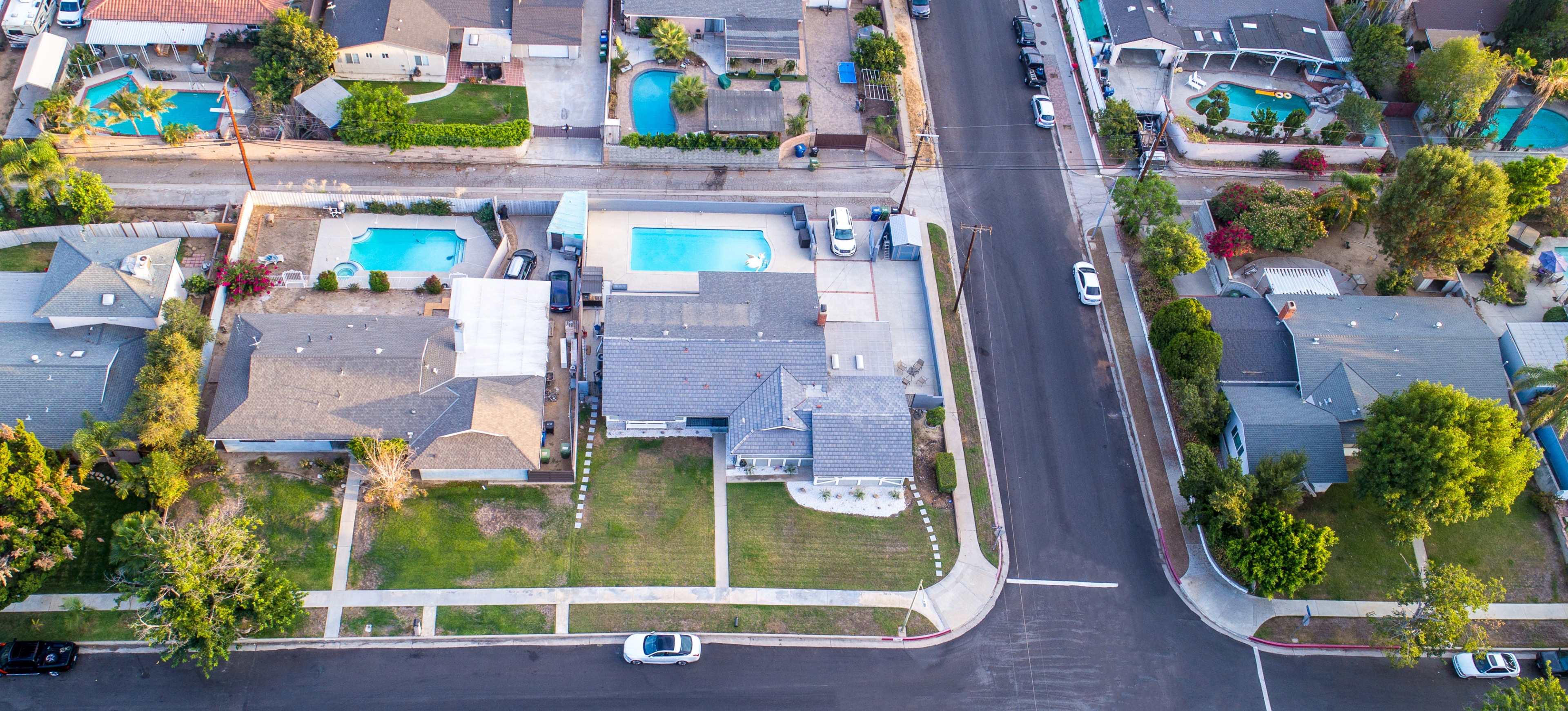 An aerial view shows a residential neighborhood with houses, swimming pools, and a paved street.
