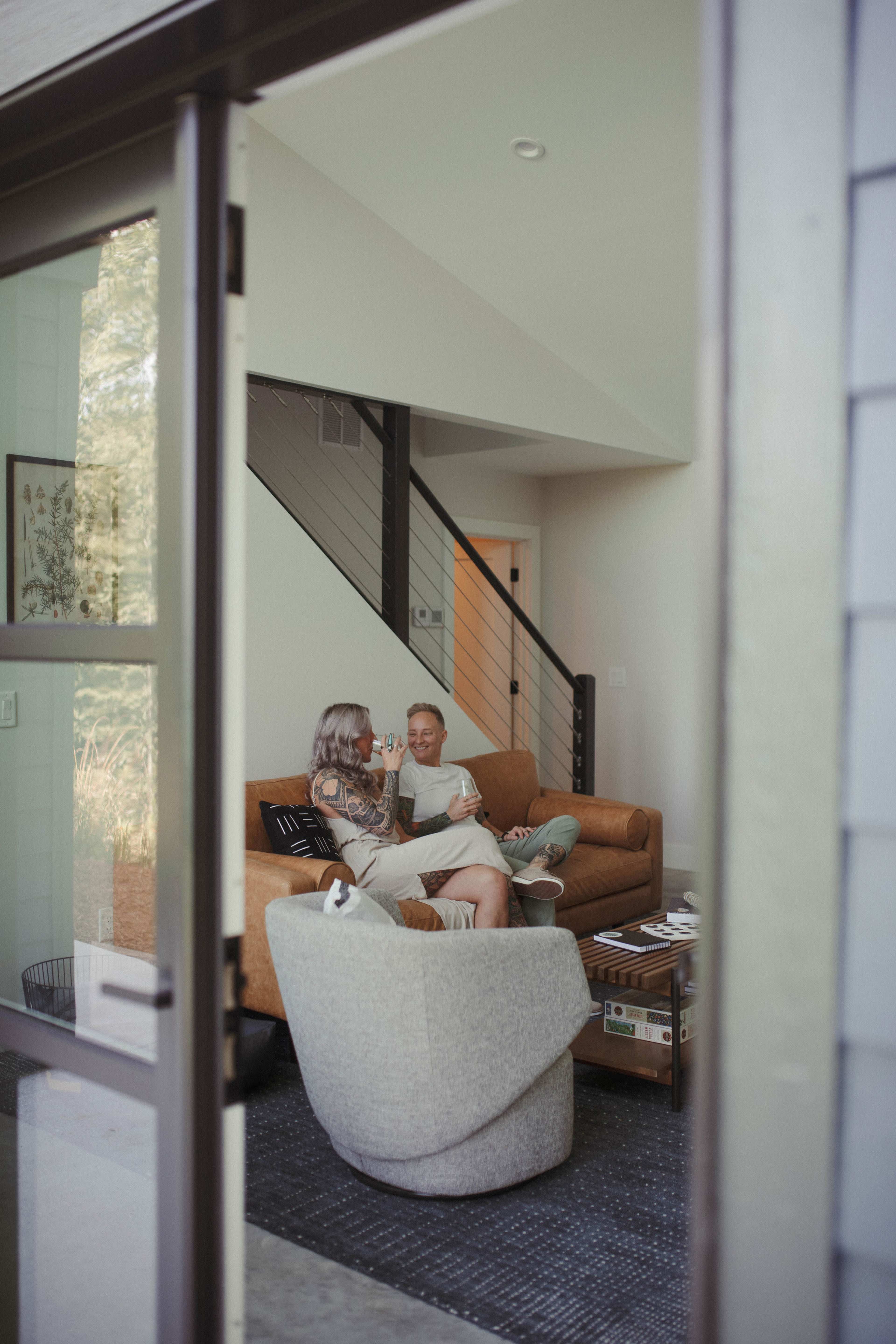 A woman and a man sit on a brown couch, enjoying drinks in a modern living room with a staircase in the background.