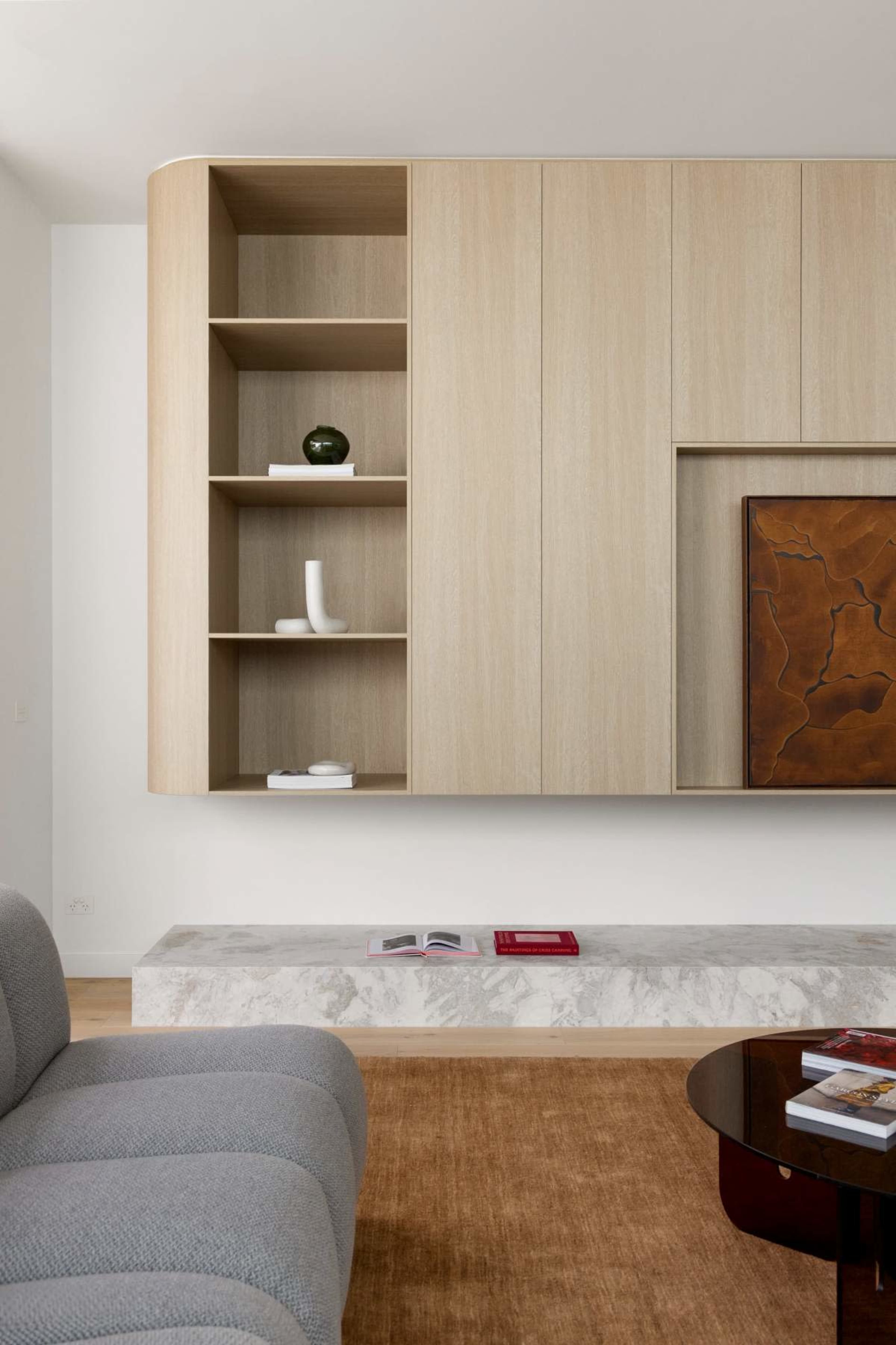 The image shows a modern interior featuring a light wooden wall unit with open shelves, a marble bench, and a round coffee table on a textured rug.