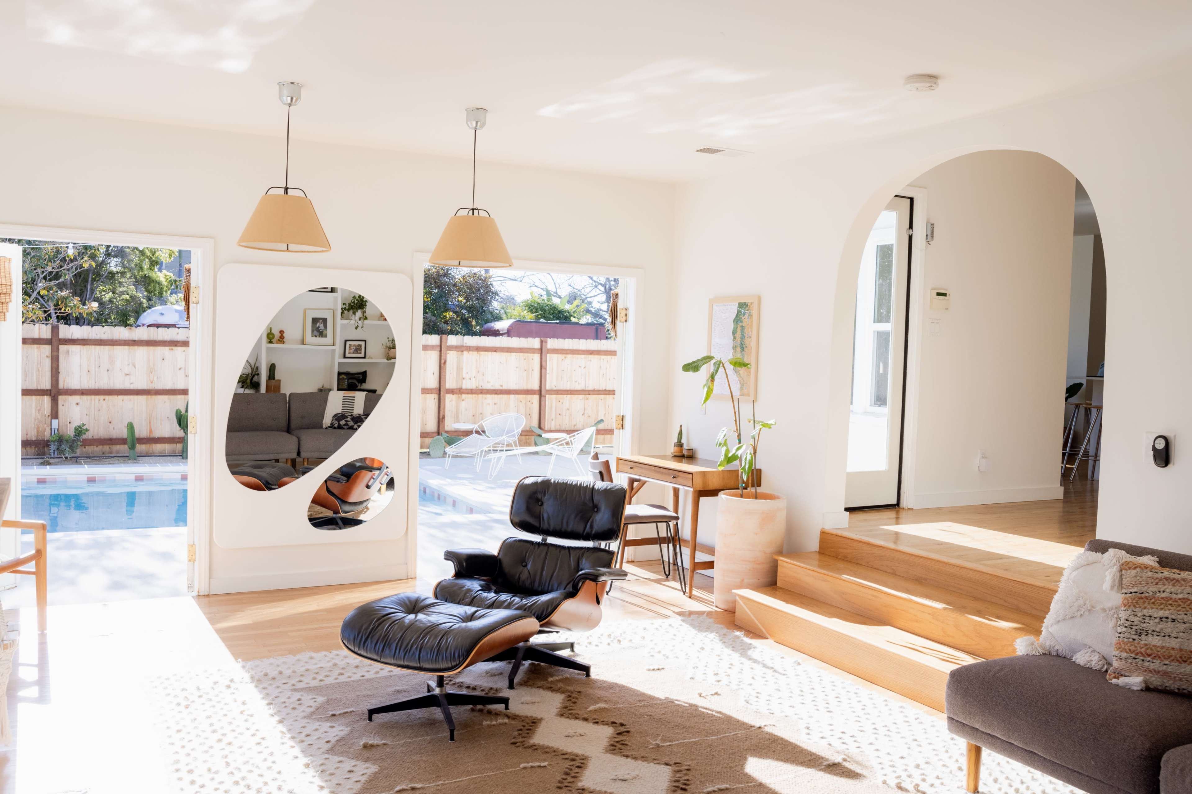 A bright, modern living room with large windows overlooking a pool area, featuring a leather lounge chair, wooden furniture, and an arched doorway leading to another room.