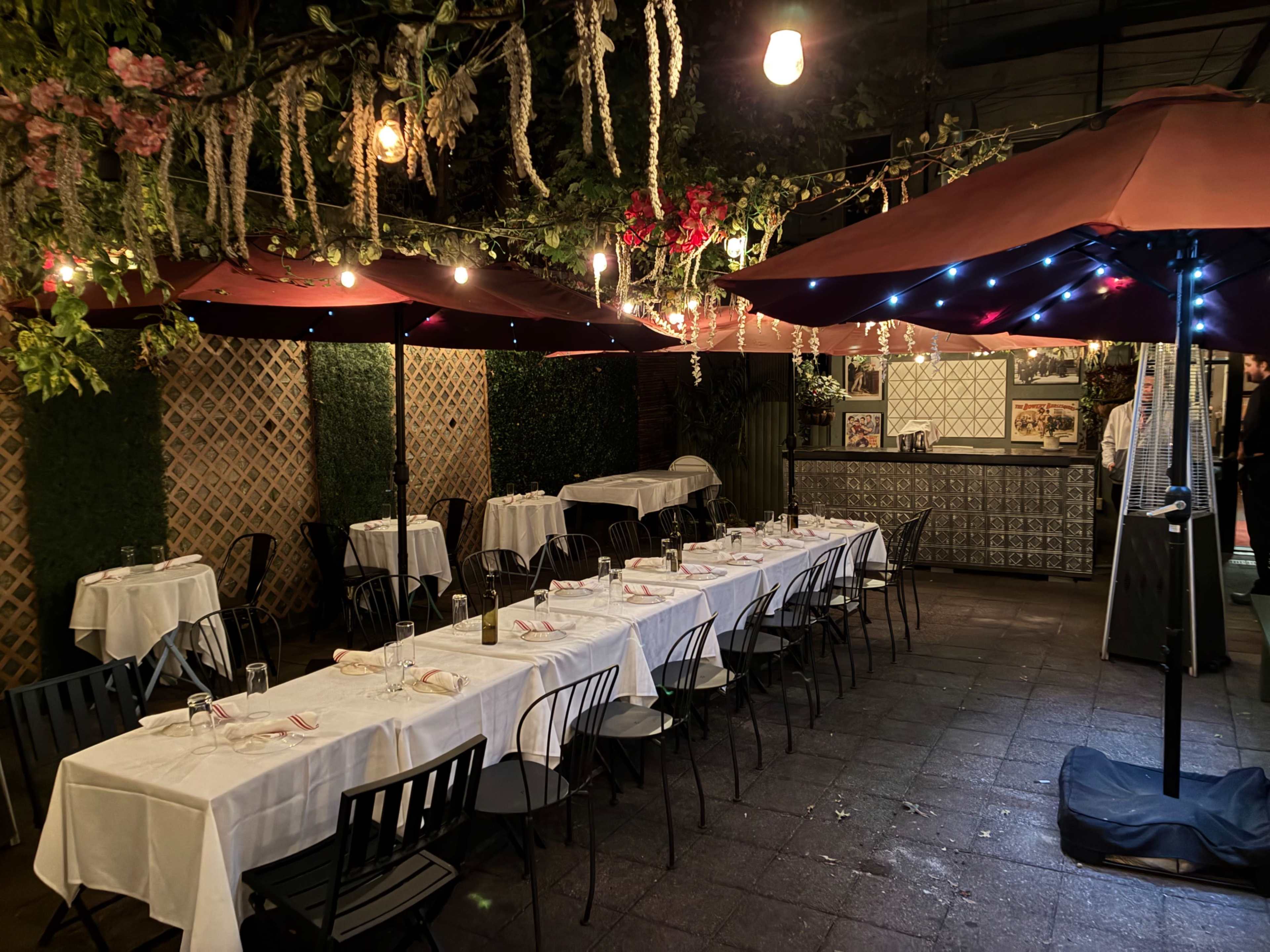 An outdoor dining area features tables covered with white tablecloths and shaded by large umbrellas, illuminated by string lights and surrounded by greenery.