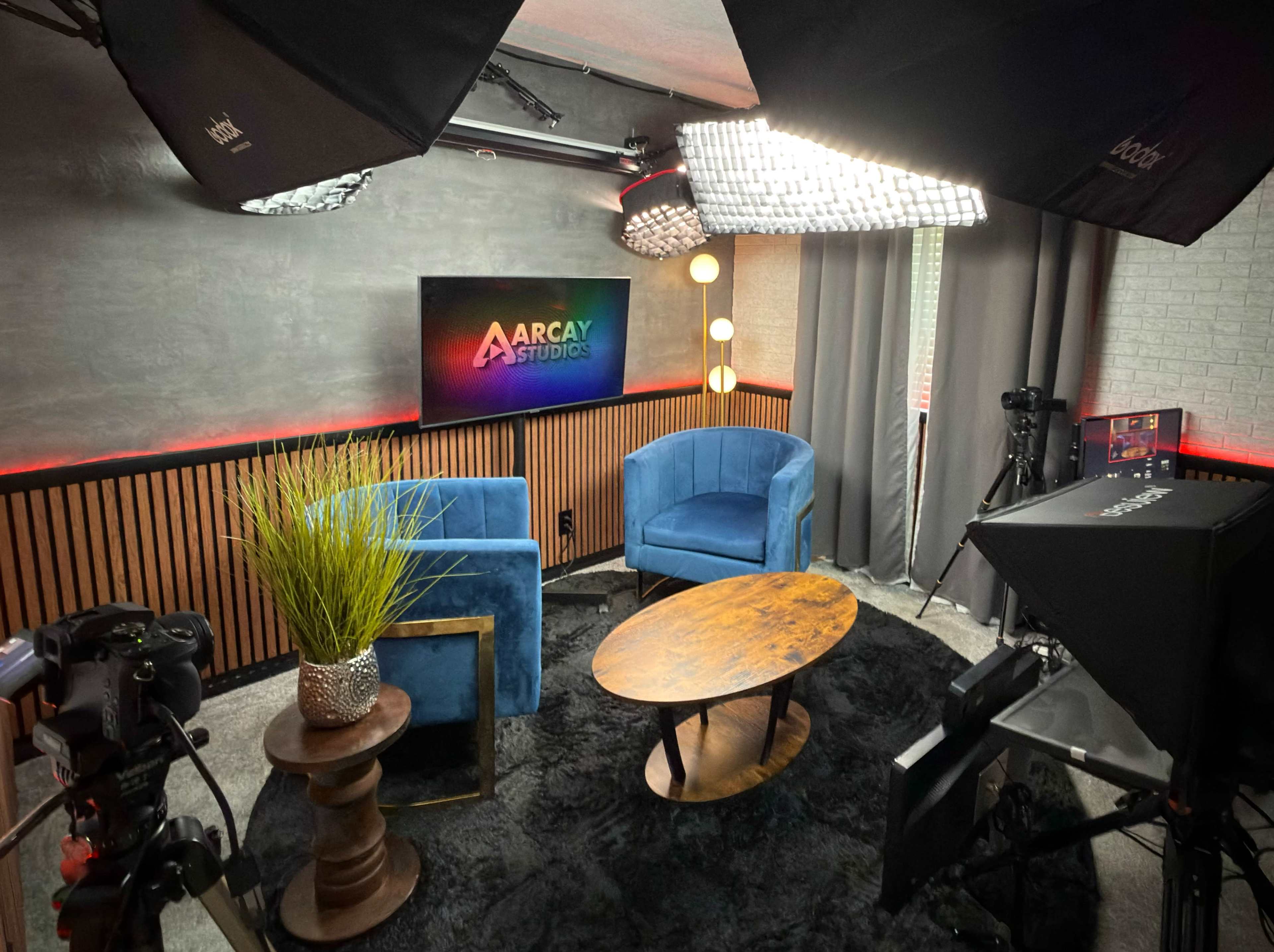 A modern studio setup features blue chairs, a wooden table, vibrant lighting, and various filming equipment.