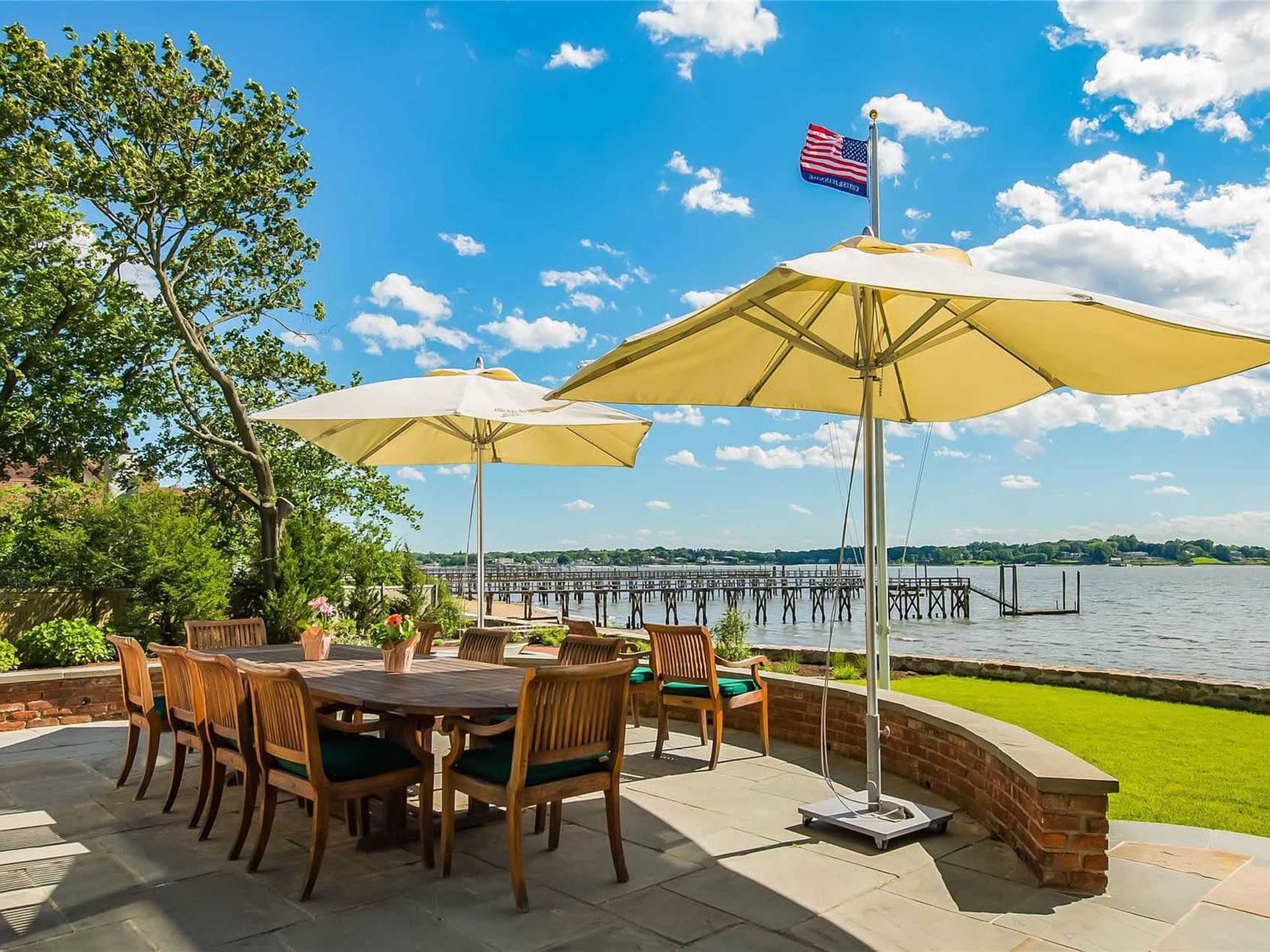The image shows a patio area with a large wooden dining table surrounded by chairs, two yellow umbrellas for shade, and a view of a river with a dock in the background.