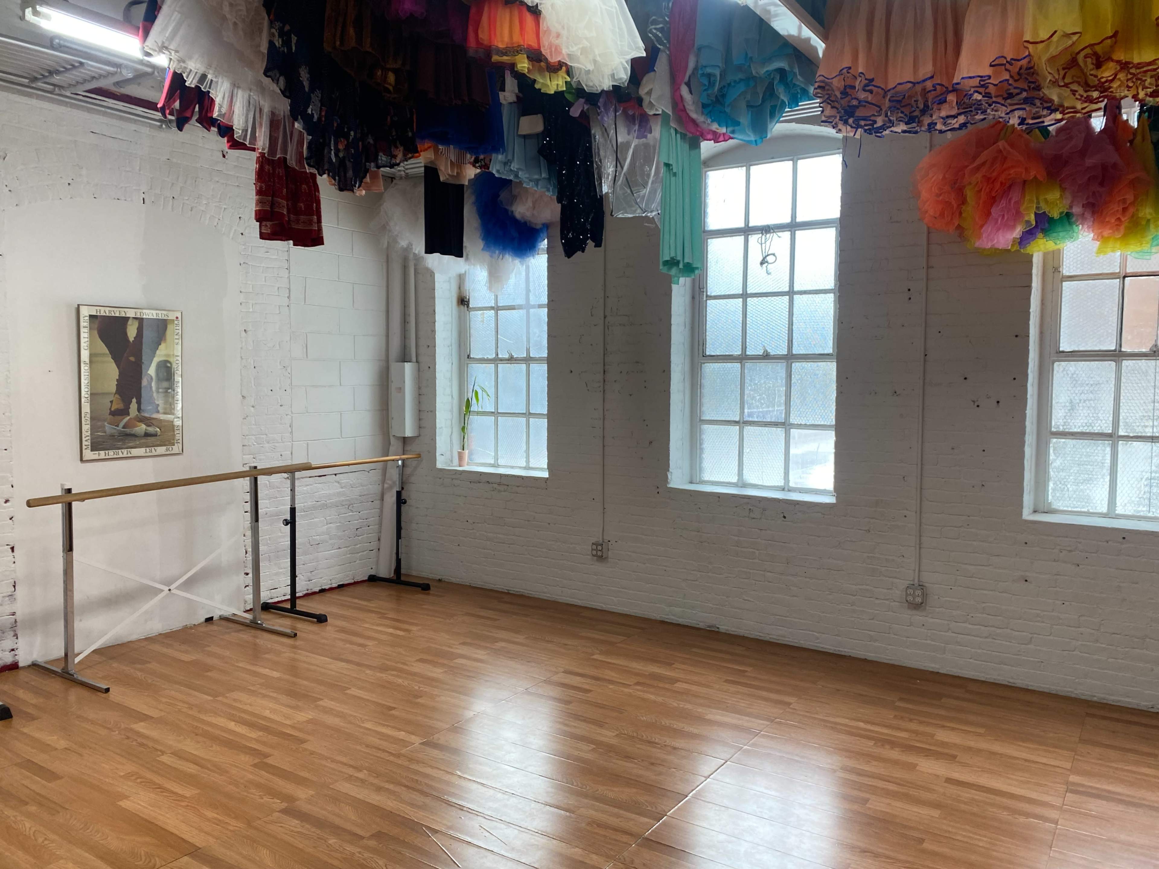 A brightly lit dance studio features wooden flooring, a ballet barre along one wall, and colorful costumes hanging from the ceiling.