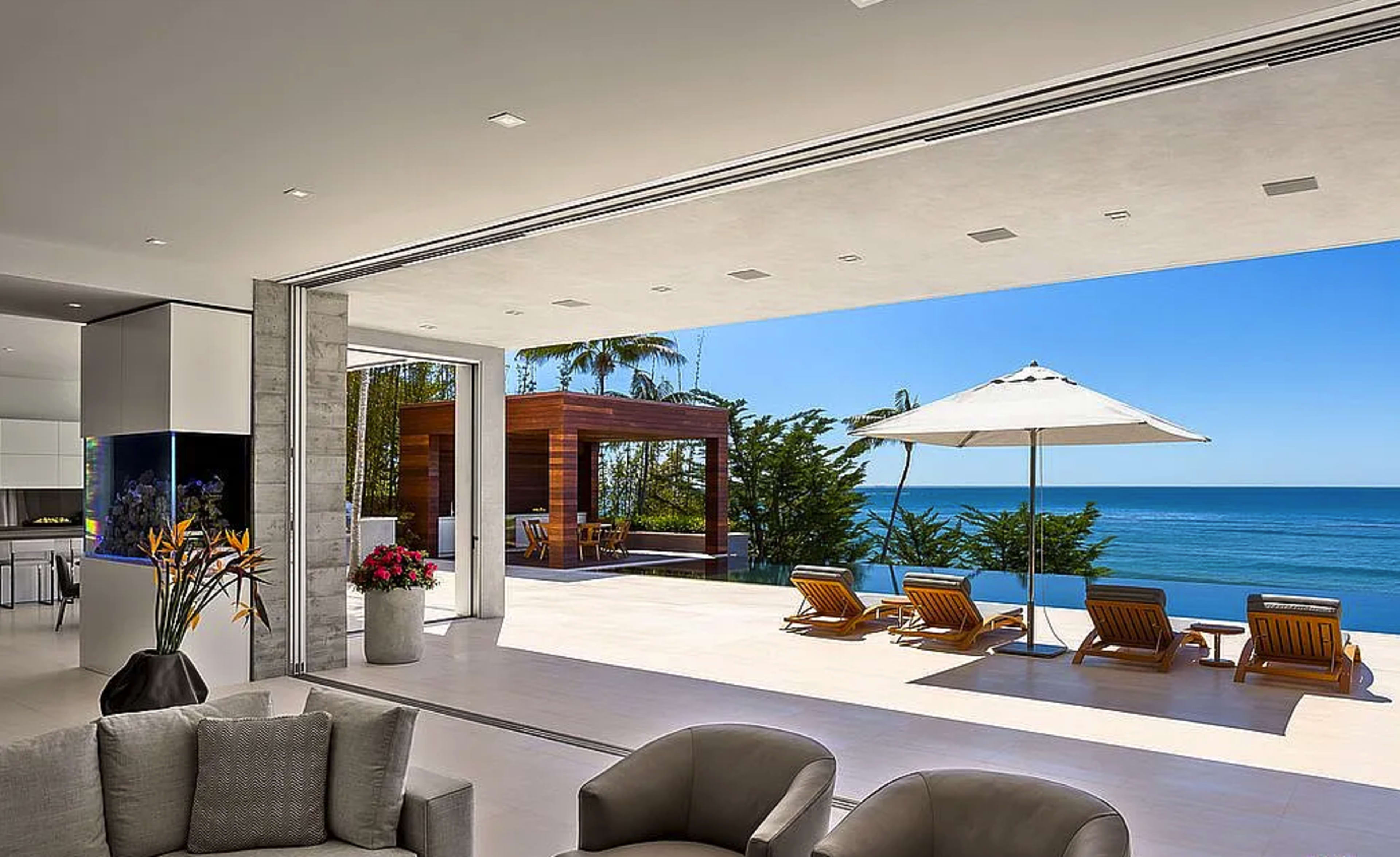 A modern living space opens to an ocean view with sliding glass doors, featuring lounge chairs and an umbrella on the patio.