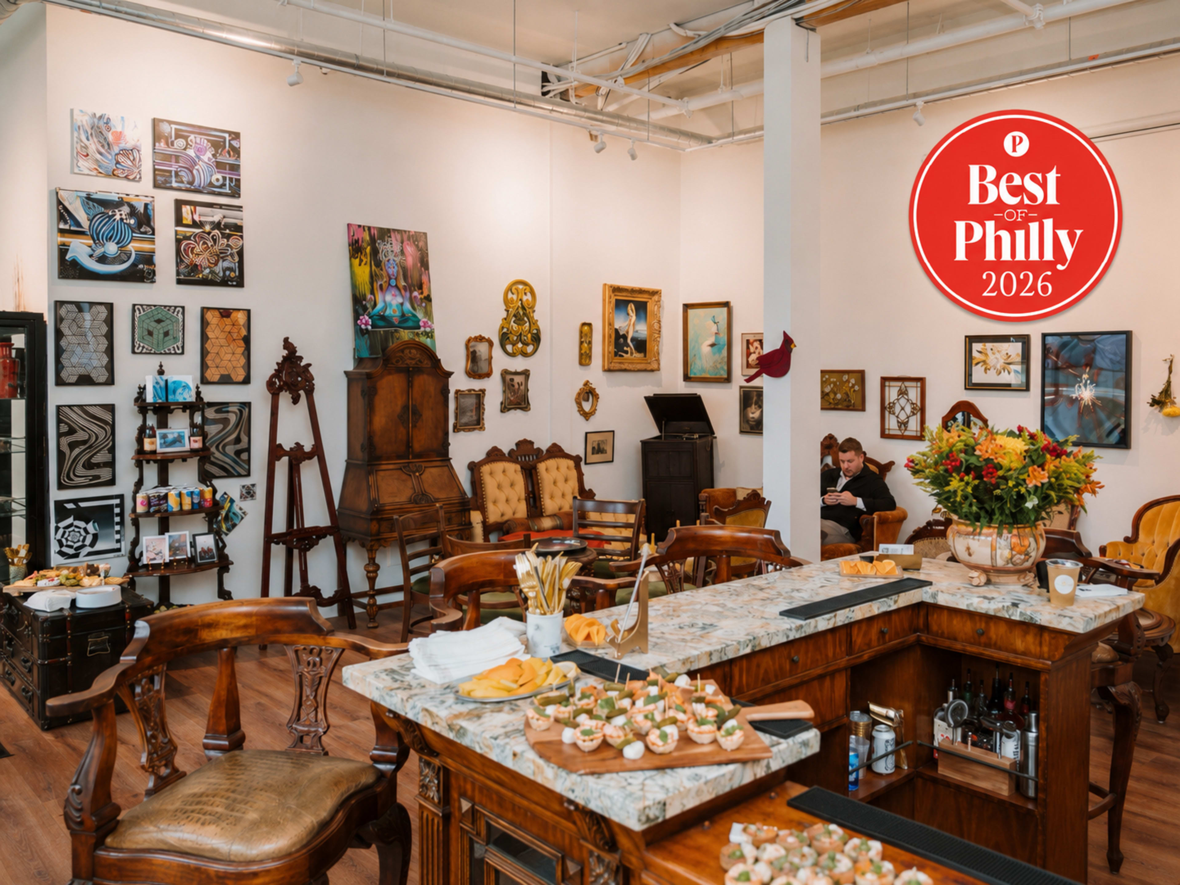 The image shows an art gallery featuring a food display on a counter, with various paintings and artworks hanging on the walls and a couple sitting together in the background.
